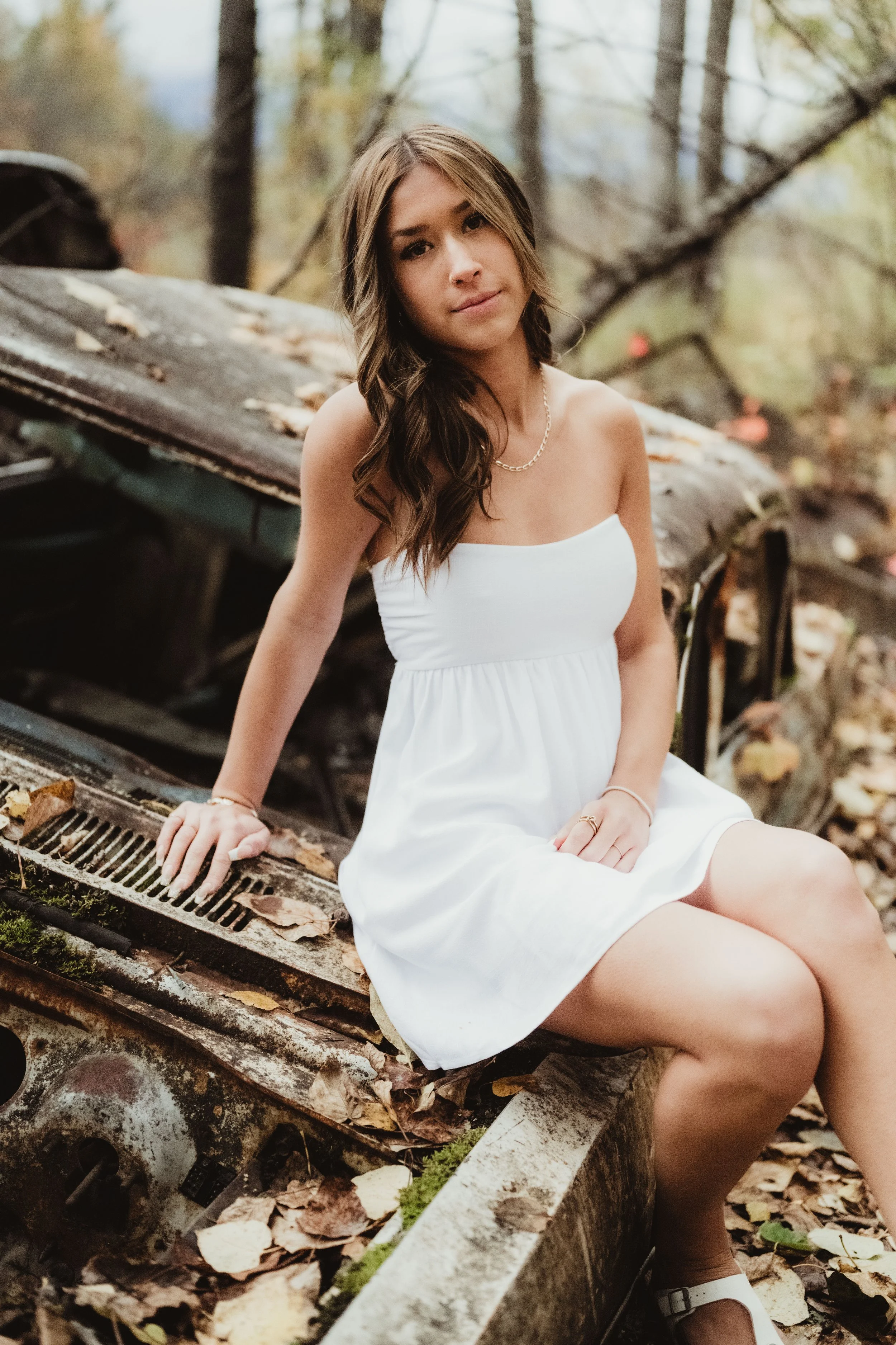 A young woman in a white dress sitting on the edge of a rusted abandoned vehicle in a forest with fallen autumn leaves.