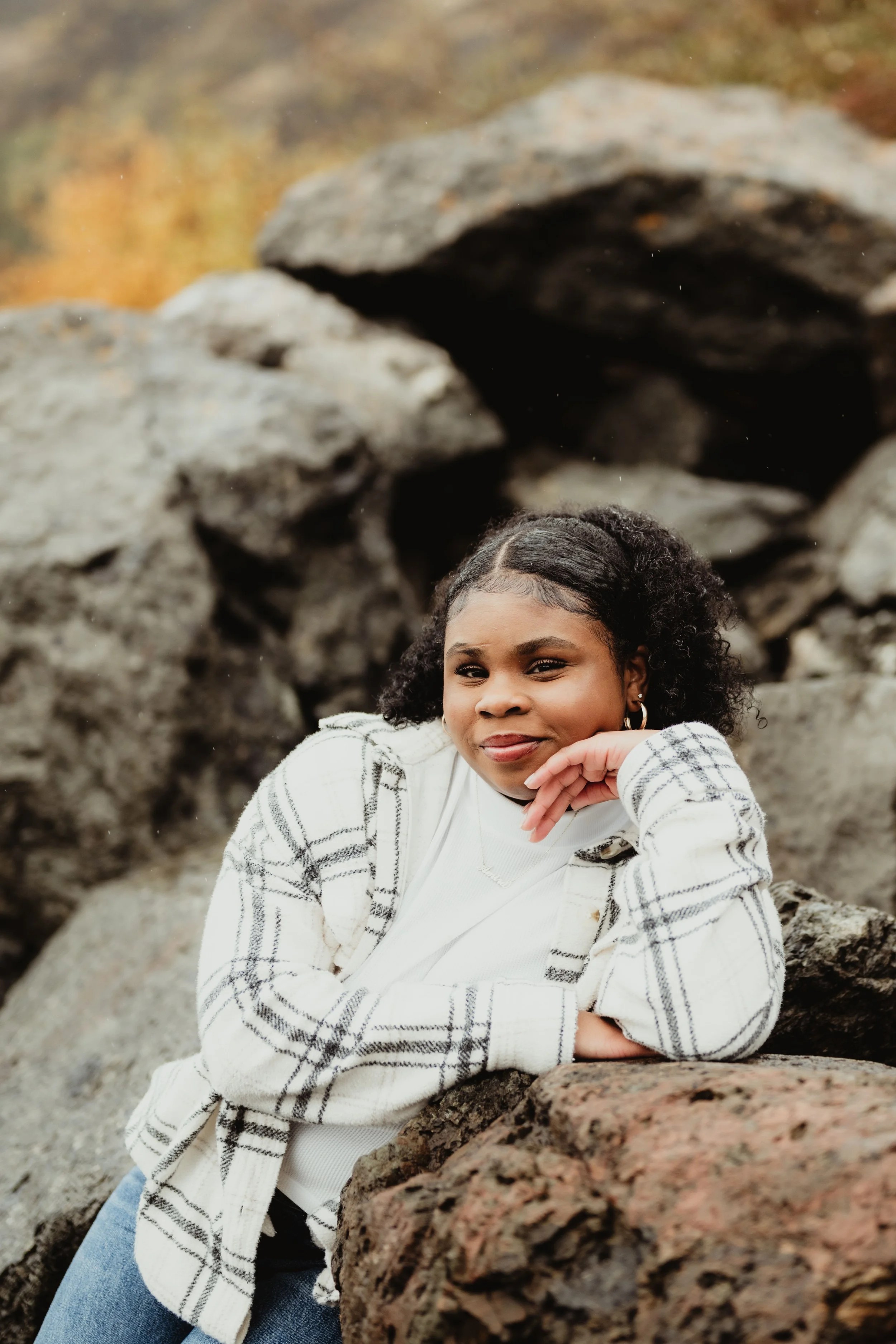 A young girl with dark curly hair and earrings, leaning on rocks outdoors, smiling at the camera with a natural background.