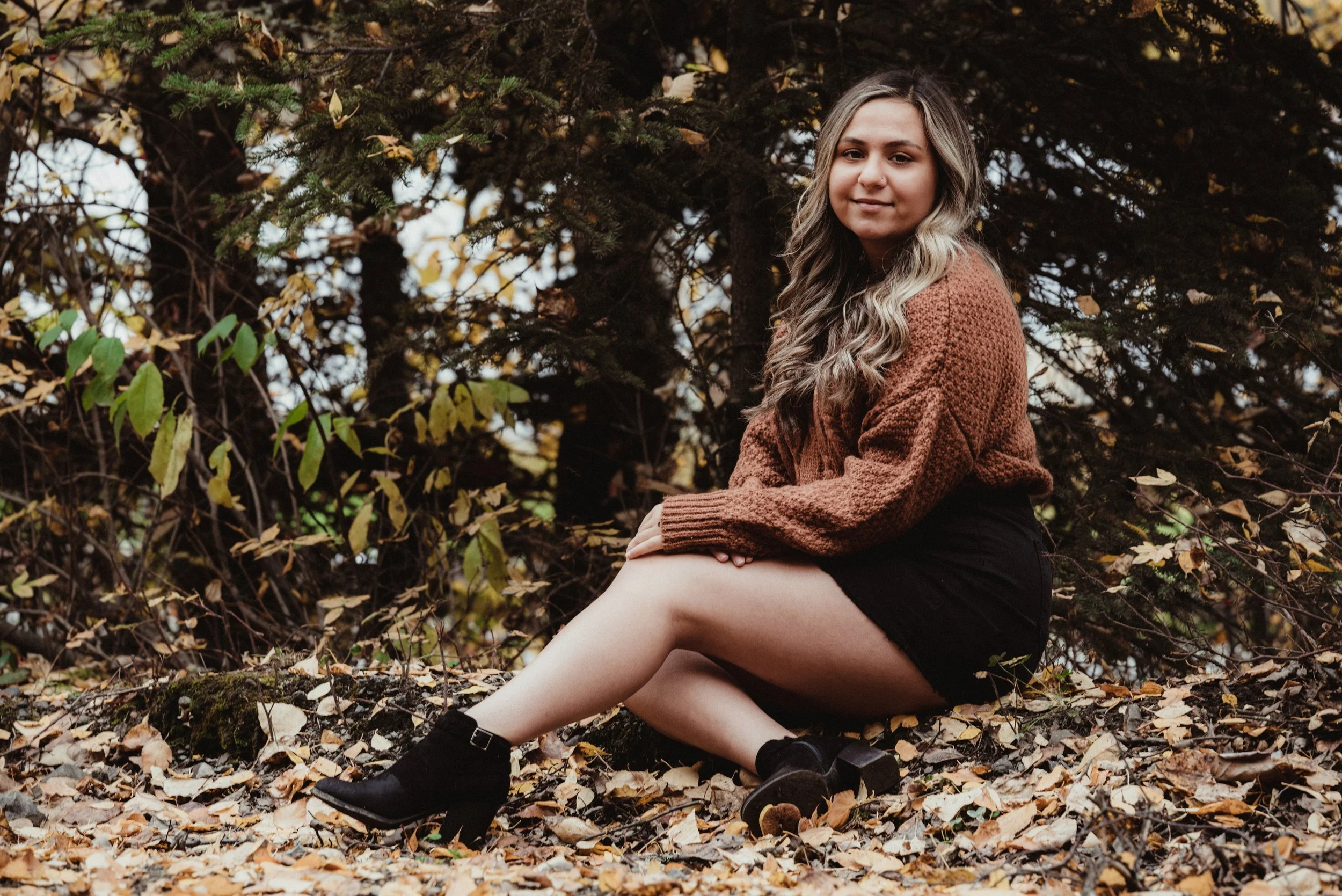 A young woman with long wavy hair sitting on a forest floor covered with fallen leaves, wearing a brown sweater, black shorts, and black ankle boots, with a background of trees and foliage.