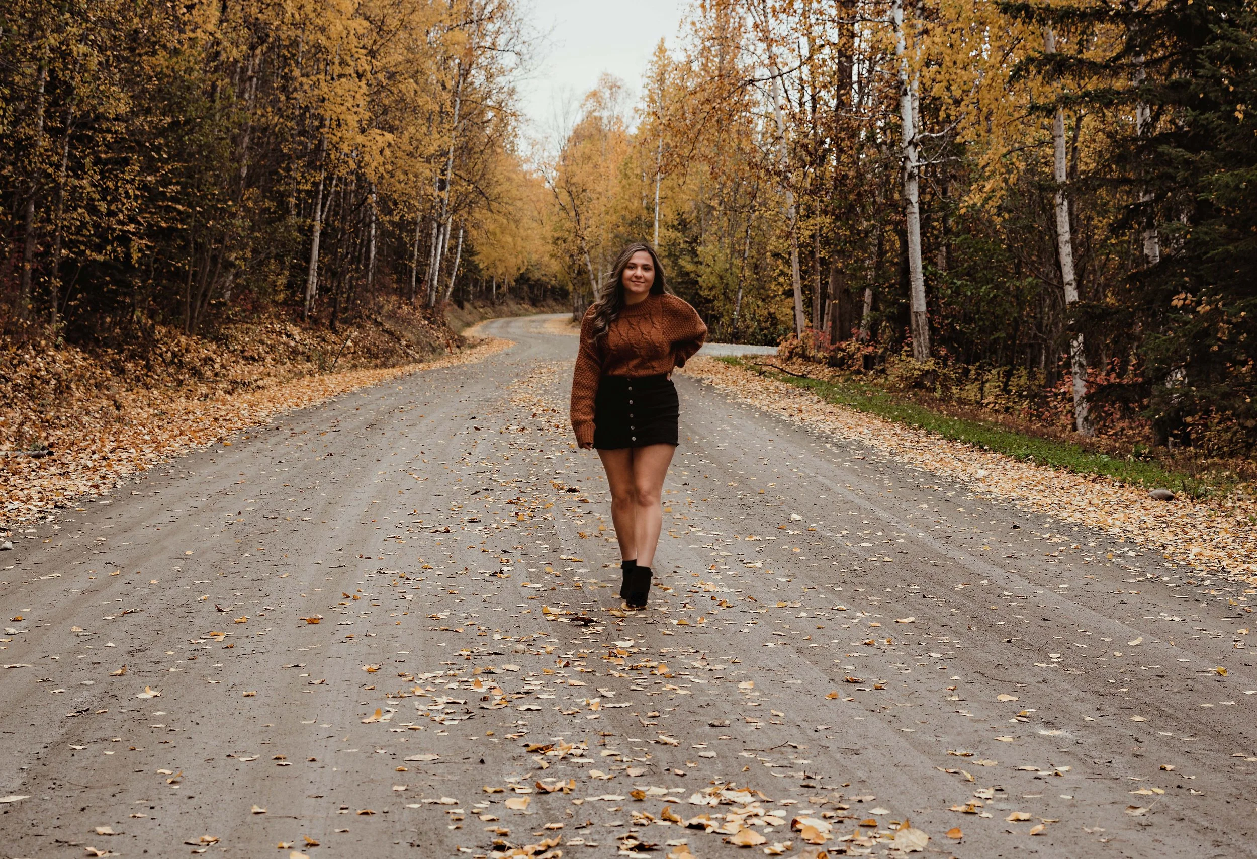 A woman walking on a dirt road surrounded by autumn trees with yellow and orange leaves.