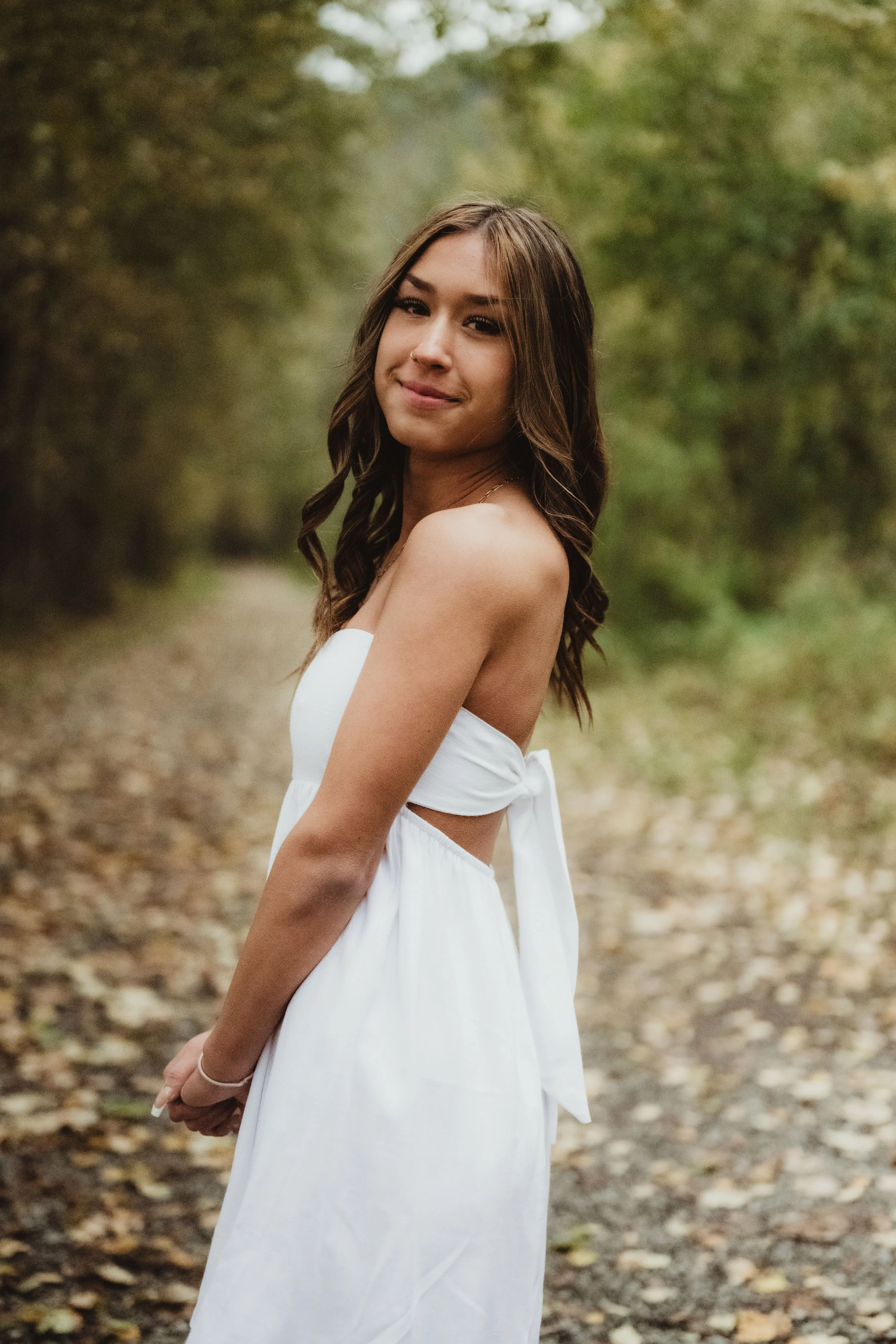 A young woman with brown wavy hair wearing a white strapless dress with a bow at the back, standing outdoors on a path surrounded by trees and fallen leaves.