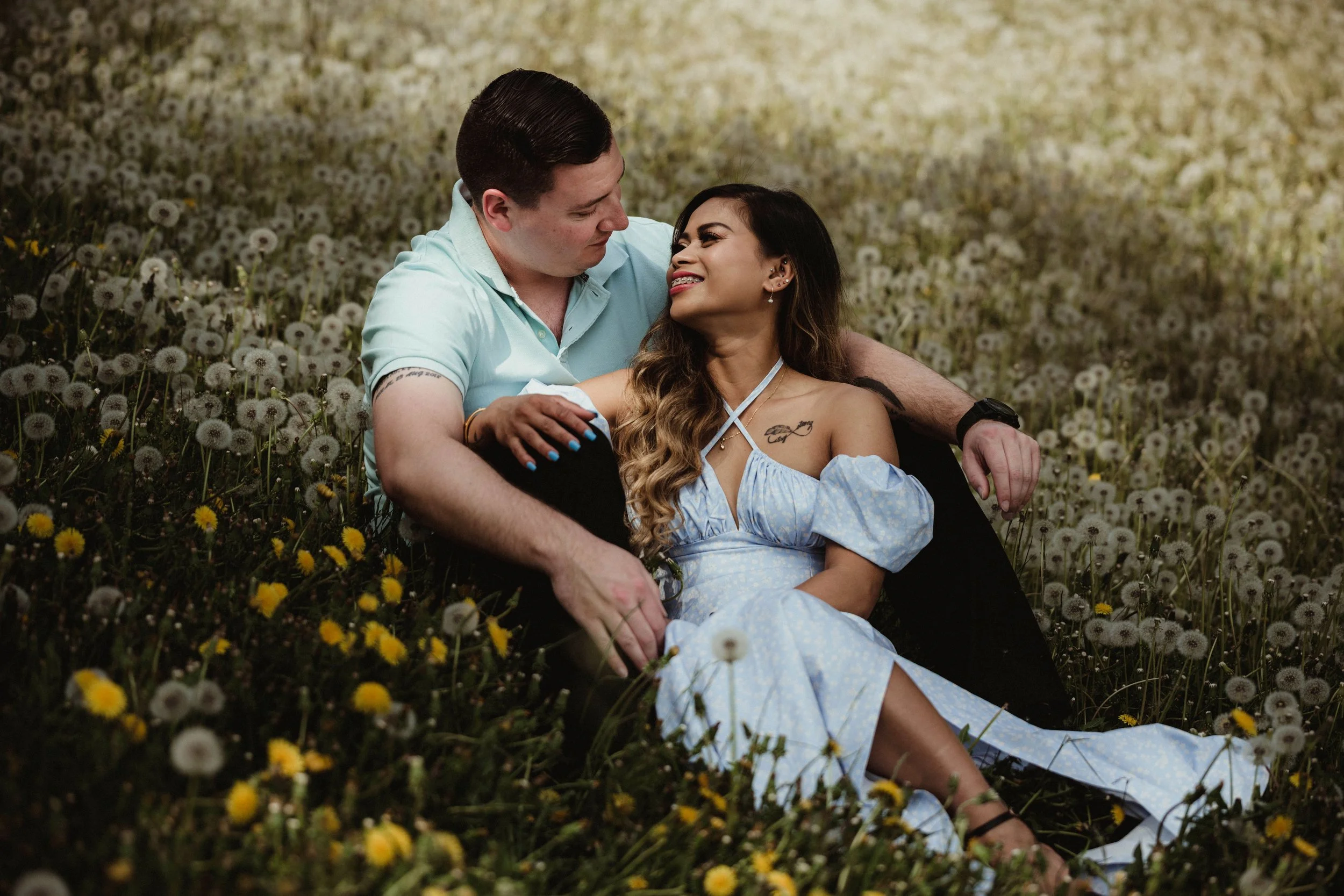 A couple sitting in a field of dandelions, gazing at each other affectionately, with the woman smiling and wearing a light blue dress.