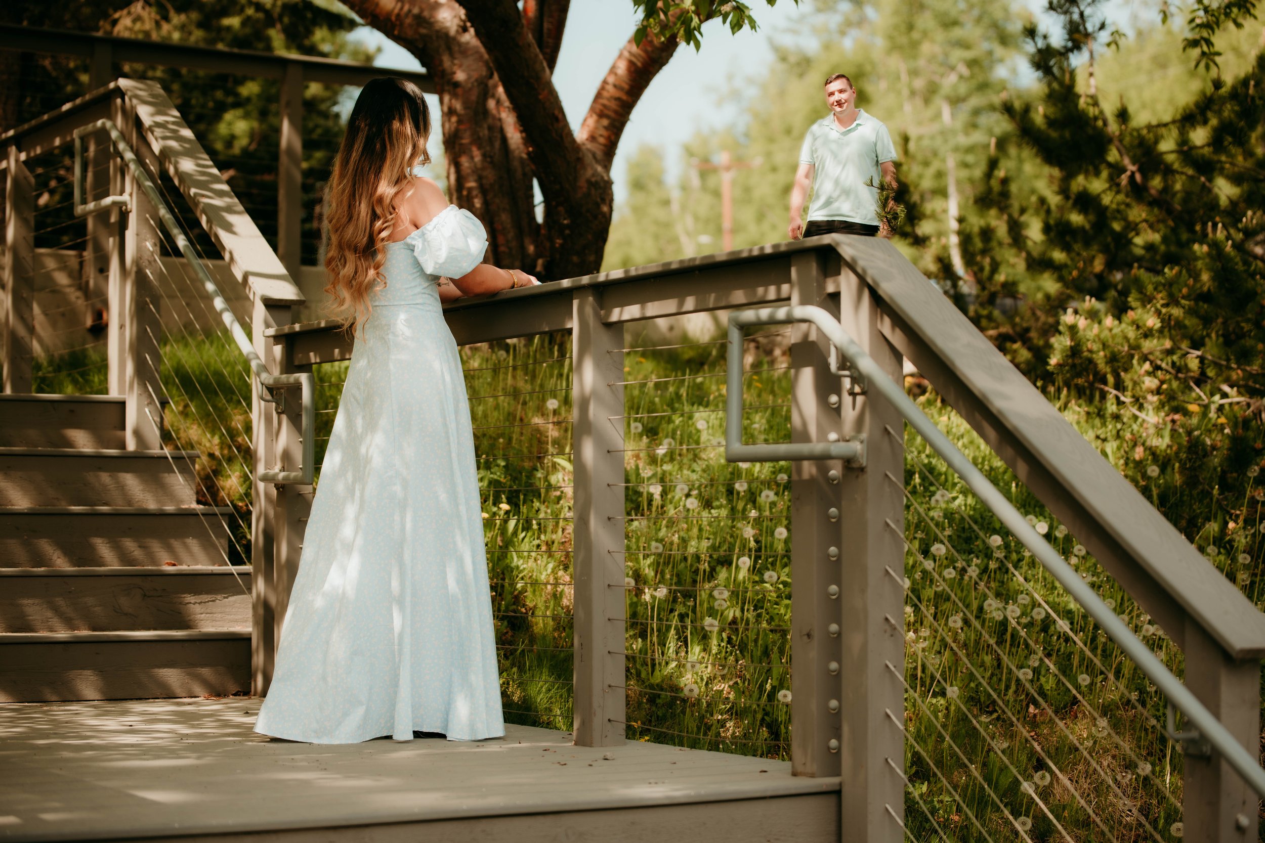 A woman in a white dress is standing on a wooden deck, looking at a man in a light green polo shirt on the ground beyond the railing, in a lush outdoor setting with trees and grass.