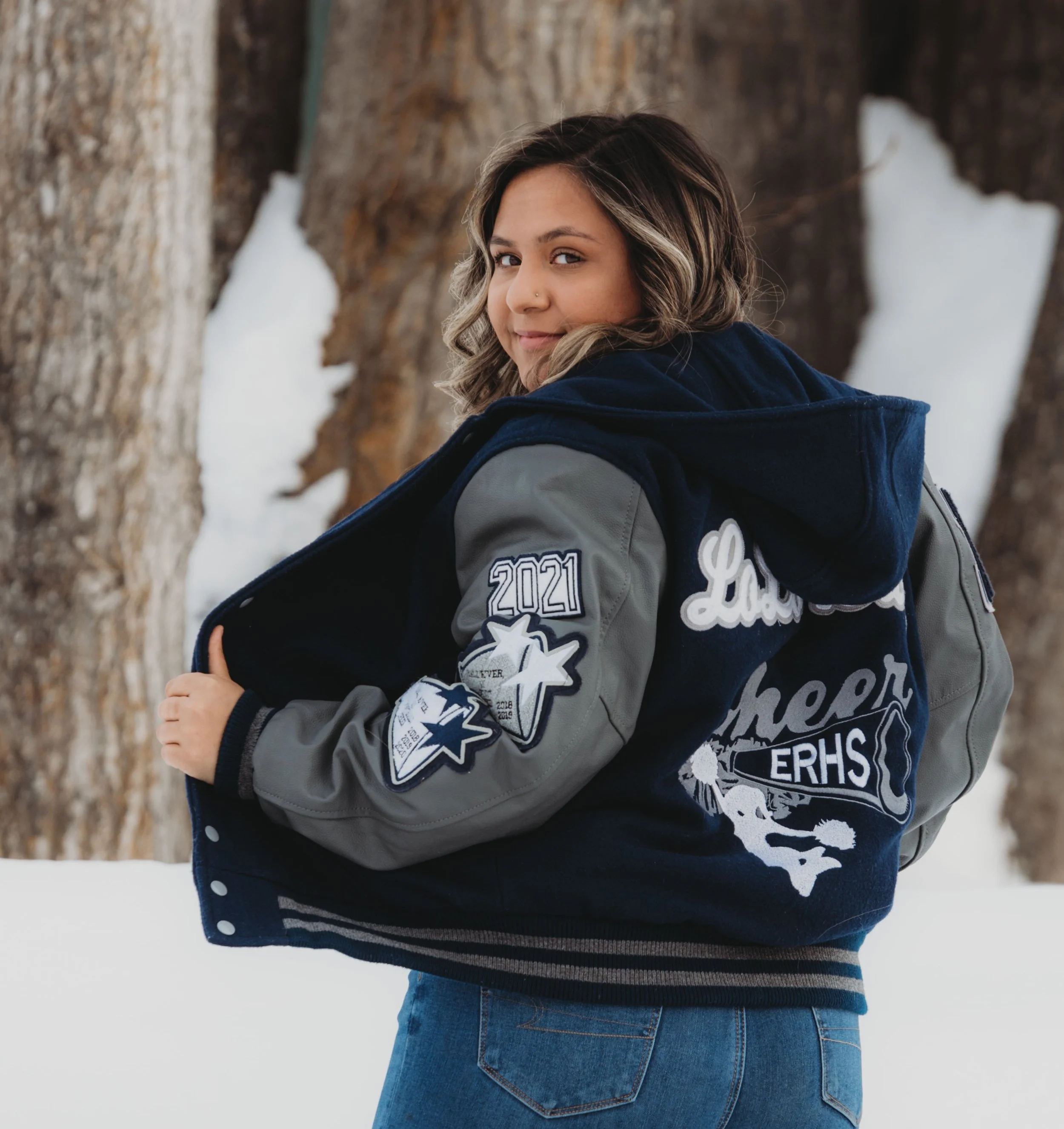 Woman wearing a varsity jacket with patches in a snowy outdoor setting, looking over her shoulder.