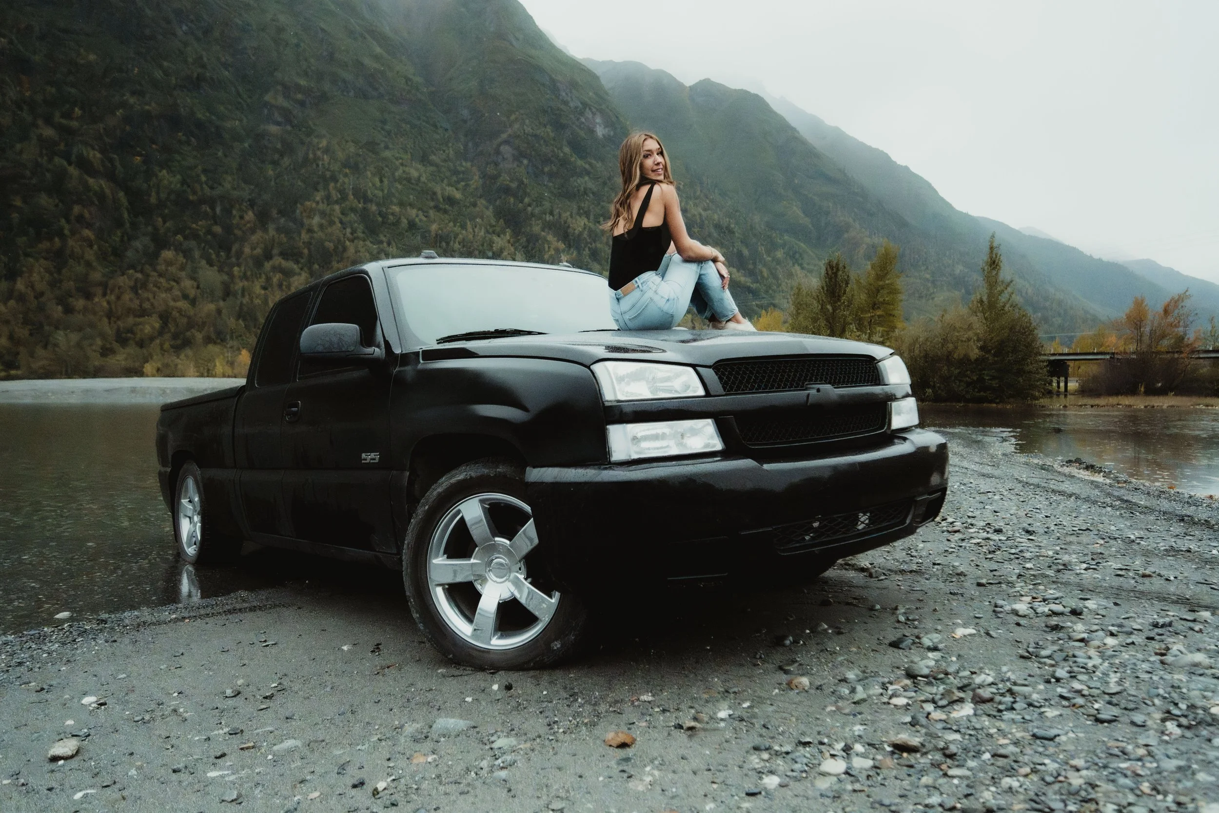 A woman sitting on the hood of a black pickup truck near a body of water, with mountains and trees in the background.