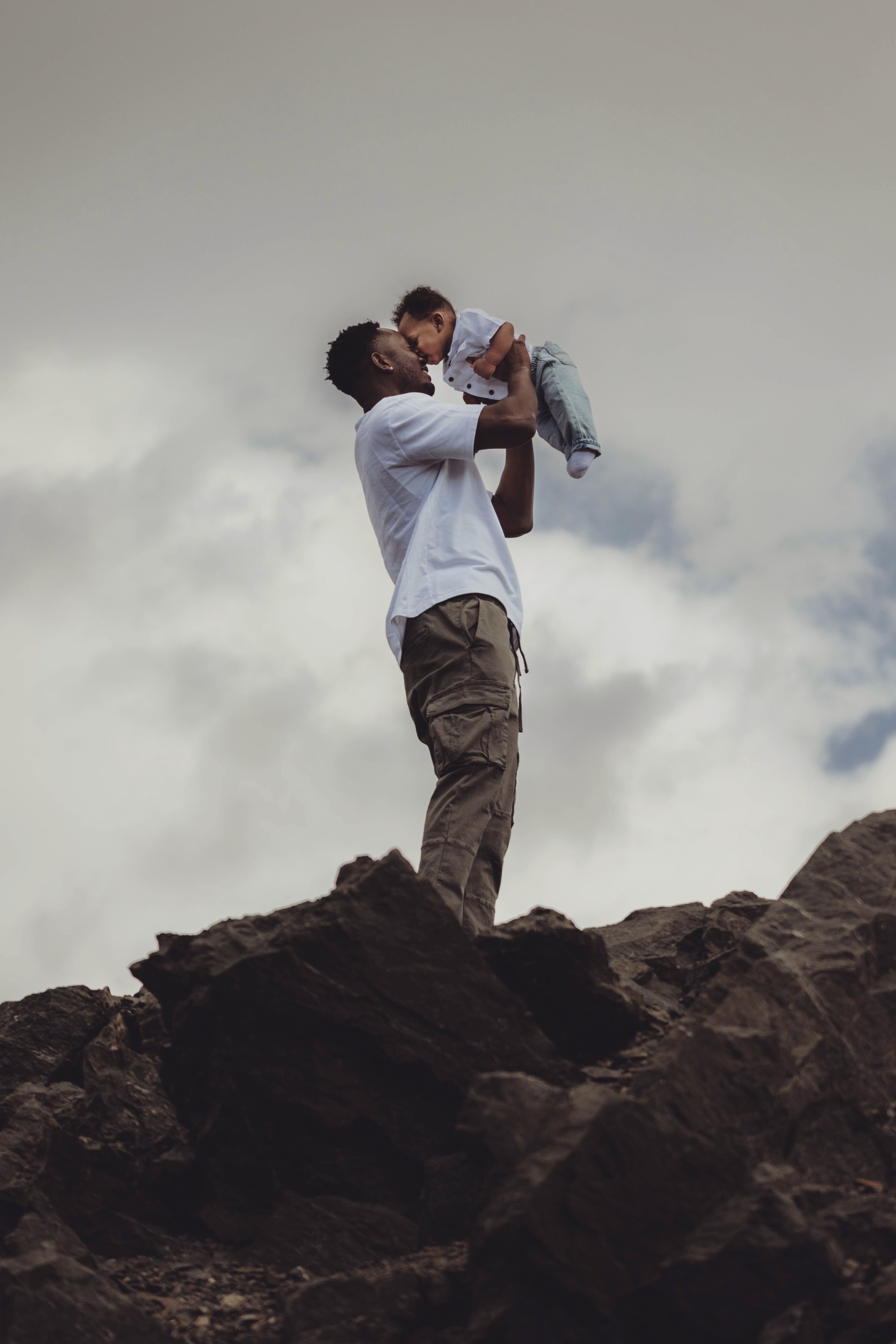 A man holding a young child up in the air on rocky terrain outdoors with cloudy sky in the background.