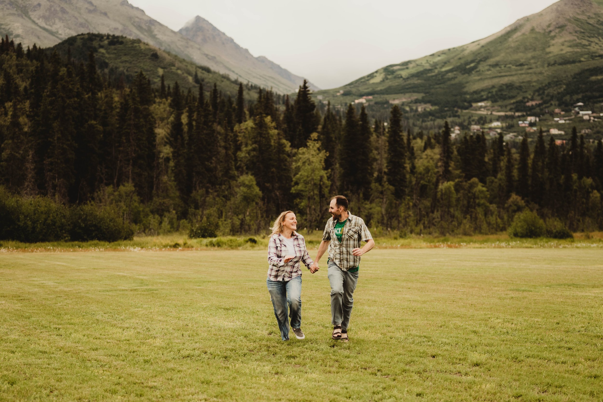 A couple walking hand in hand on a grassy field with mountains and a dense forest in the background.