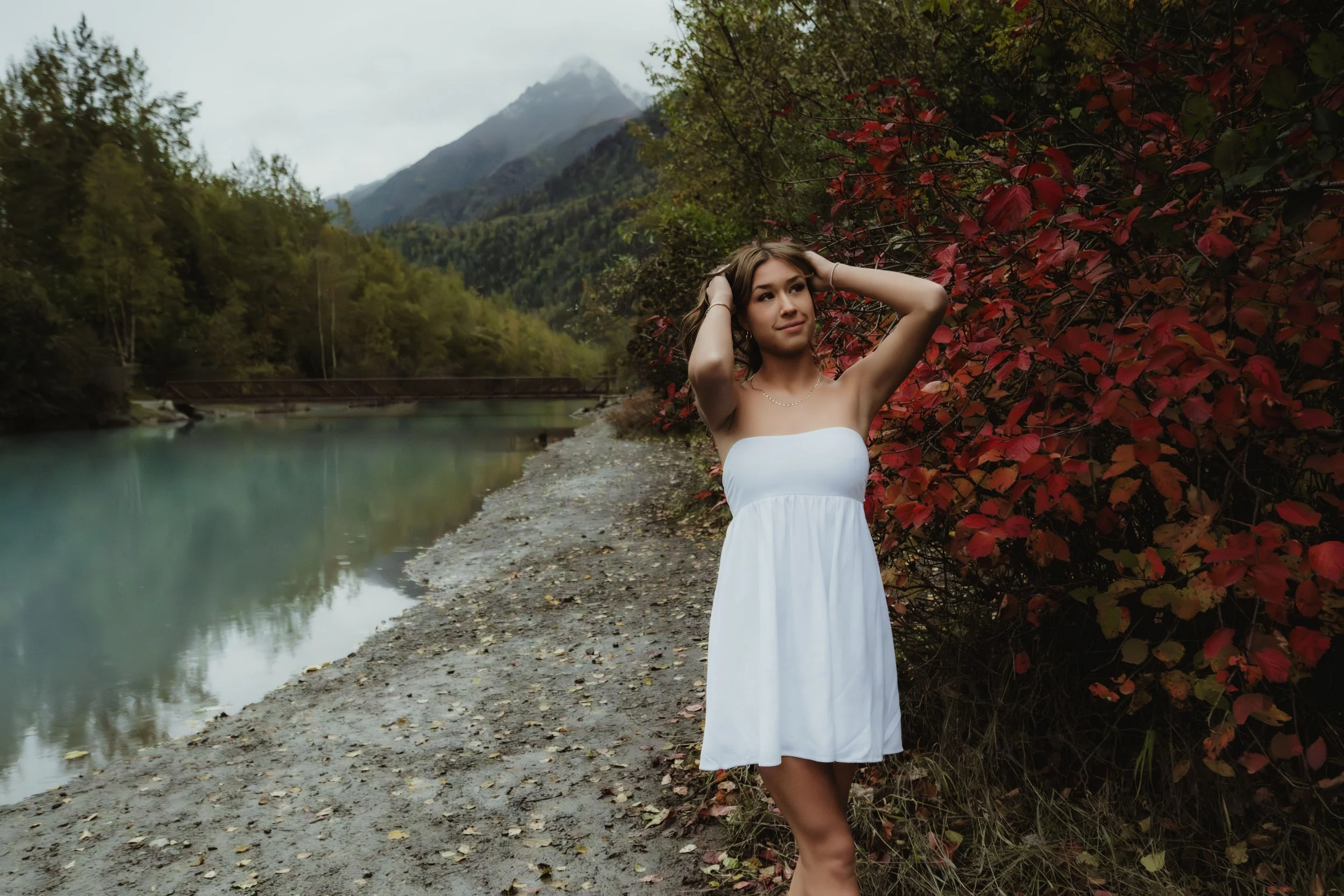 Young woman in a white strapless dress standing near red foliage by a mountain lake with forested trees and mountain peaks in the background.