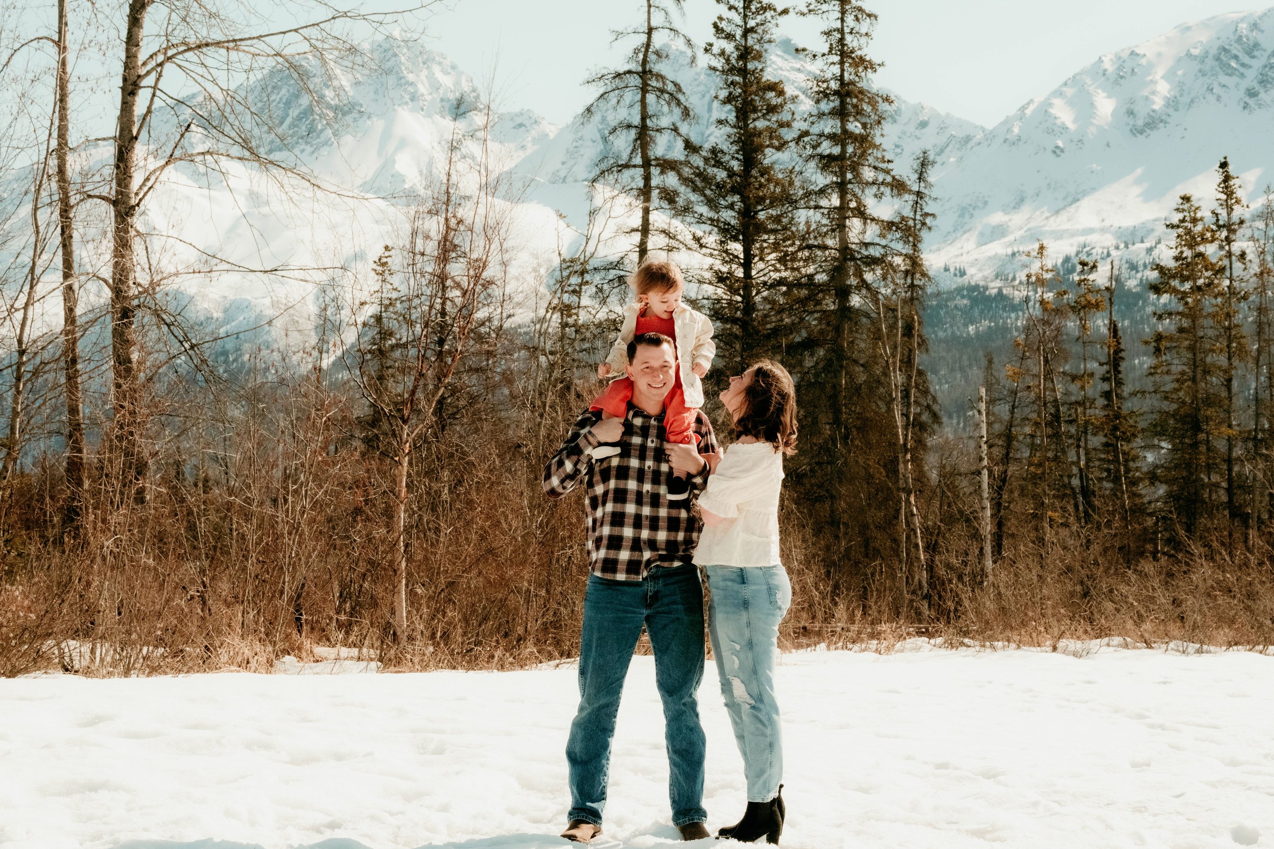 Family of three enjoying snow in a mountain landscape, with snow-covered peaks and tall trees in the background.