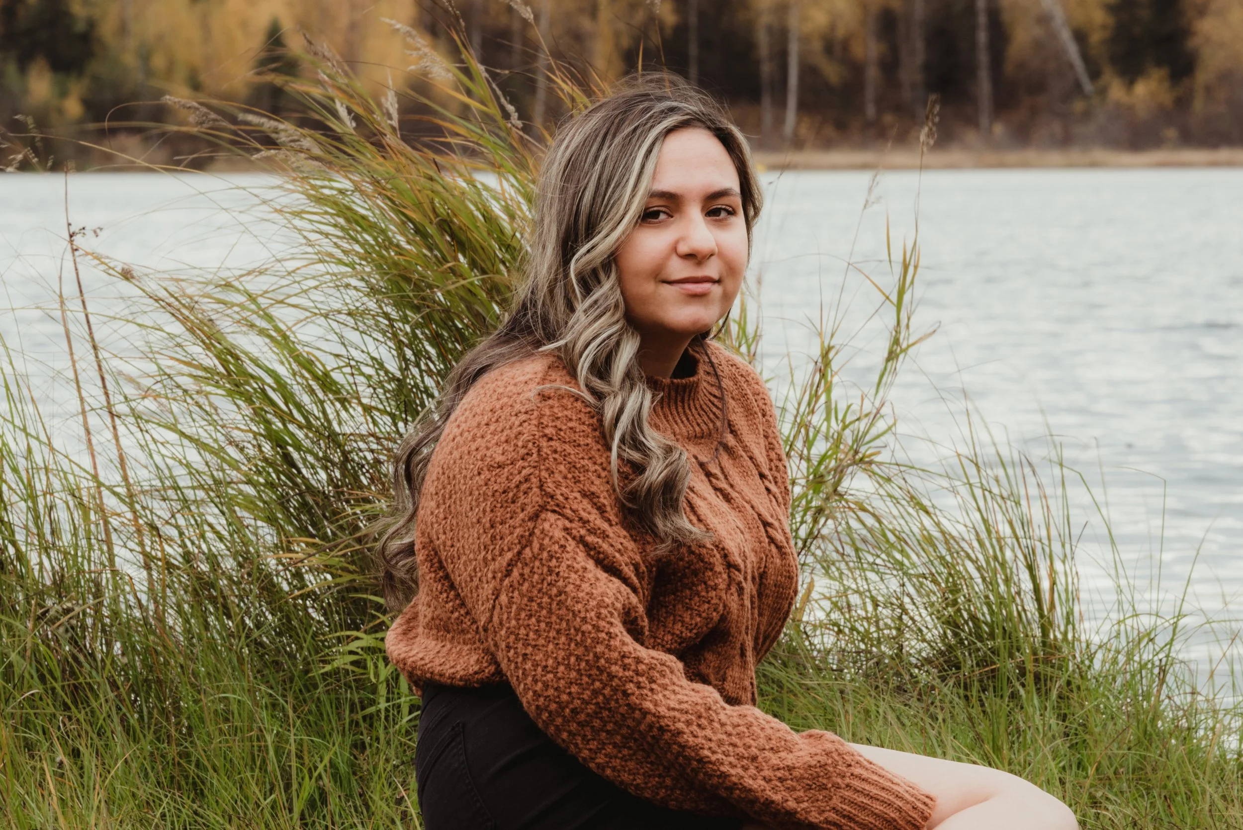 Young woman with long wavy hair sitting by a body of water, surrounded by tall grass, during autumn.