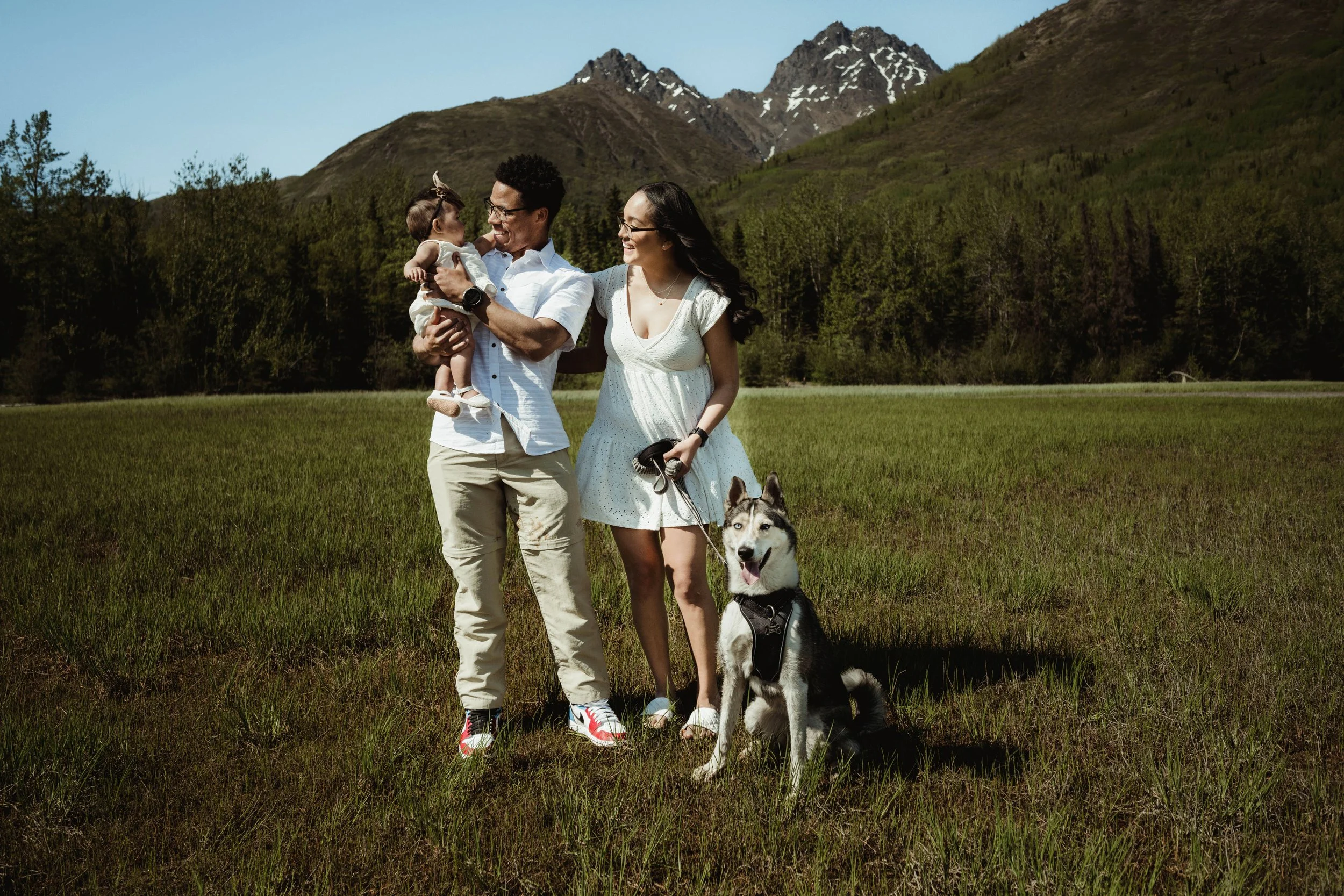 A family of three with a dog standing in a grassy field with mountains in the background. The father is holding a baby girl, and the mother is holding a leash attached to a Siberian Husky. The family is smiling and enjoying a sunny day outdoors.