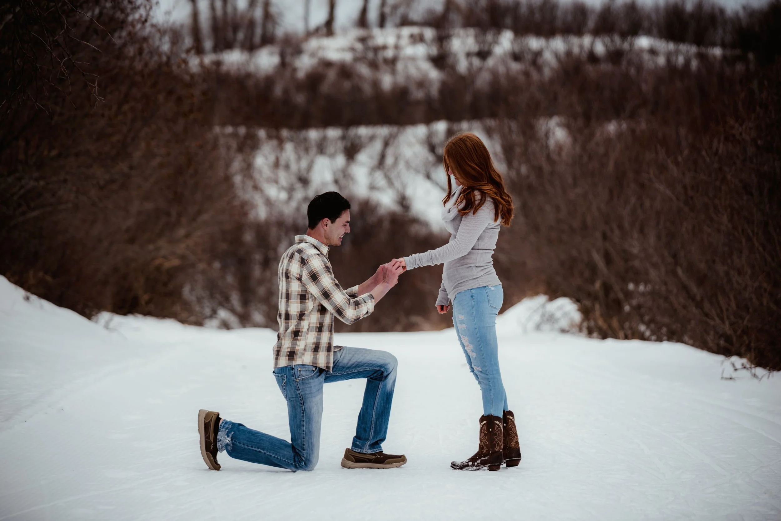 A man proposes to a woman on a snowy pathway during winter, with trees in the background.