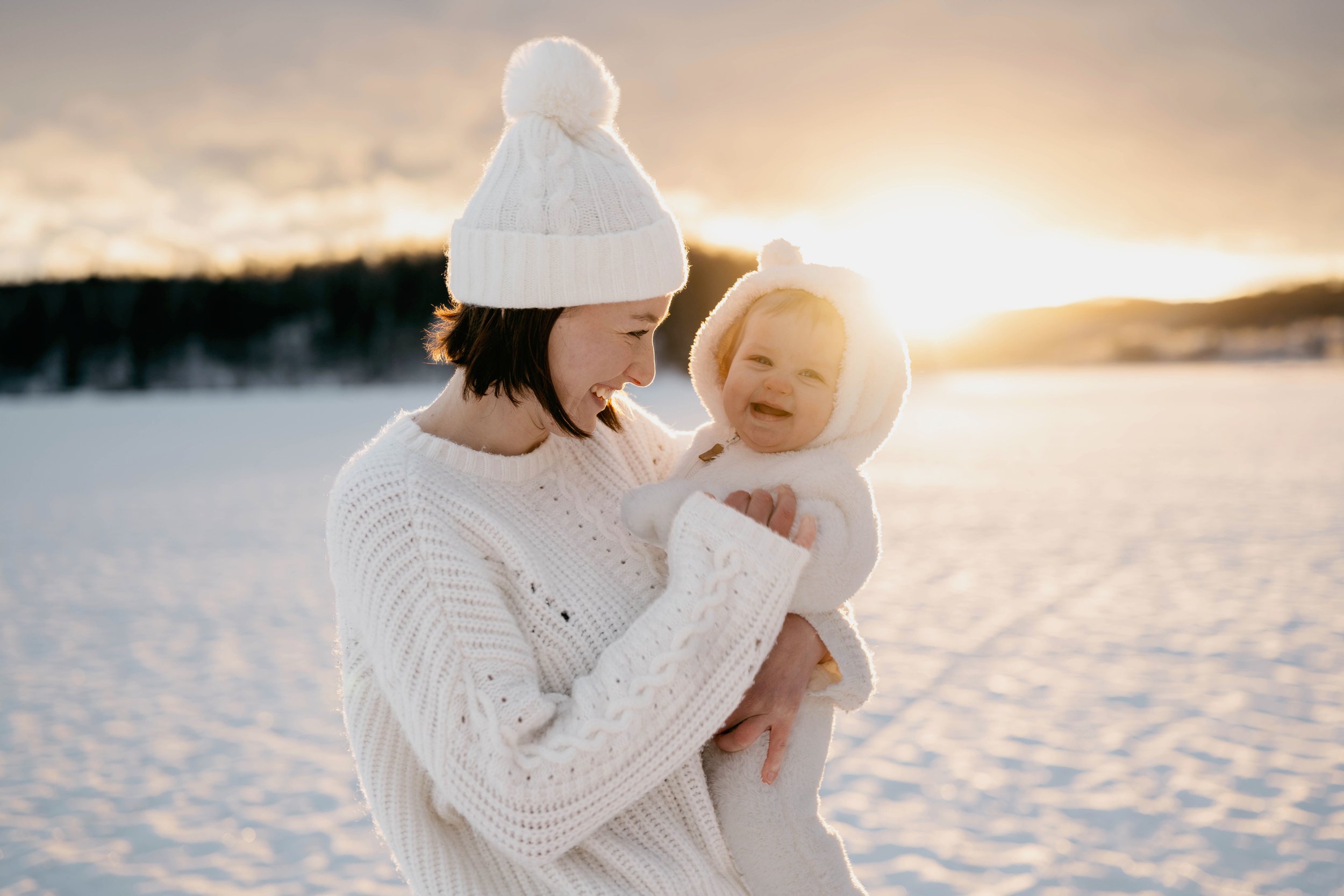 A woman and a baby dressed in white winter clothing, standing in a snowy landscape at sunset, smiling and enjoying each other's company.