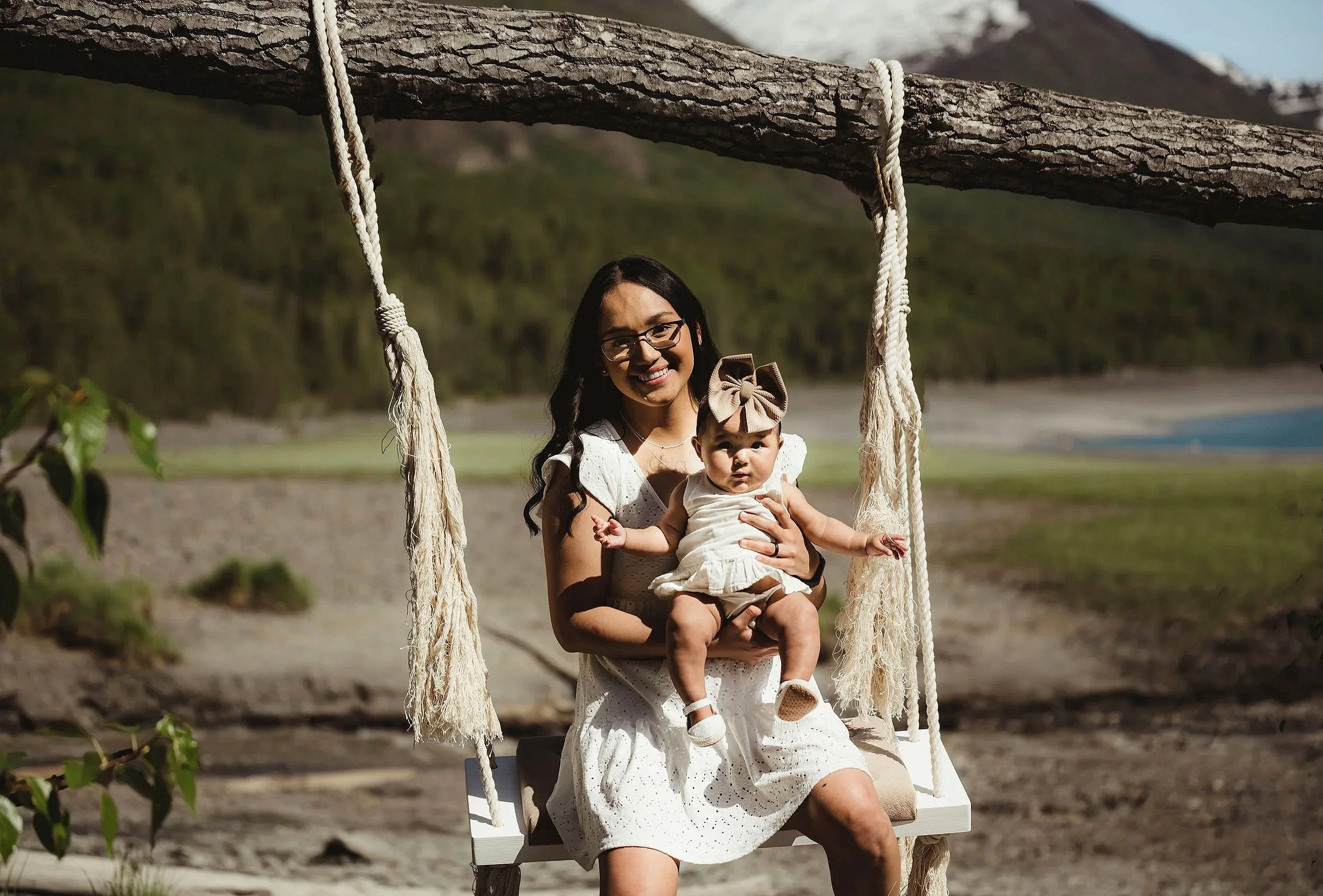 A woman and a baby girl sitting on a swing outdoors with a grassy landscape, trees, mountains, and a body of water in the background, both smiling.