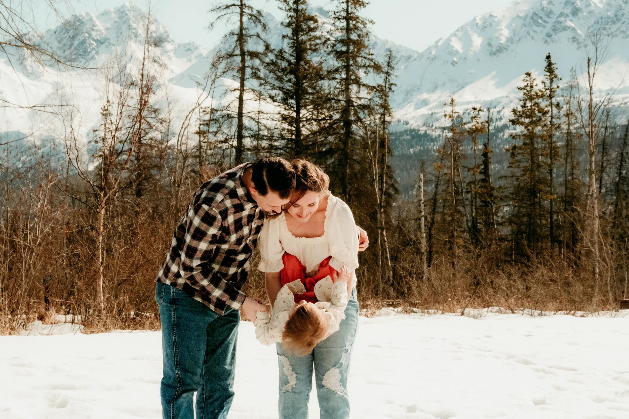A family of three enjoying a snowy outdoor setting with mountains and trees in the background. The parents are holding their young daughter upside down, all smiling and wearing casual winter clothes.