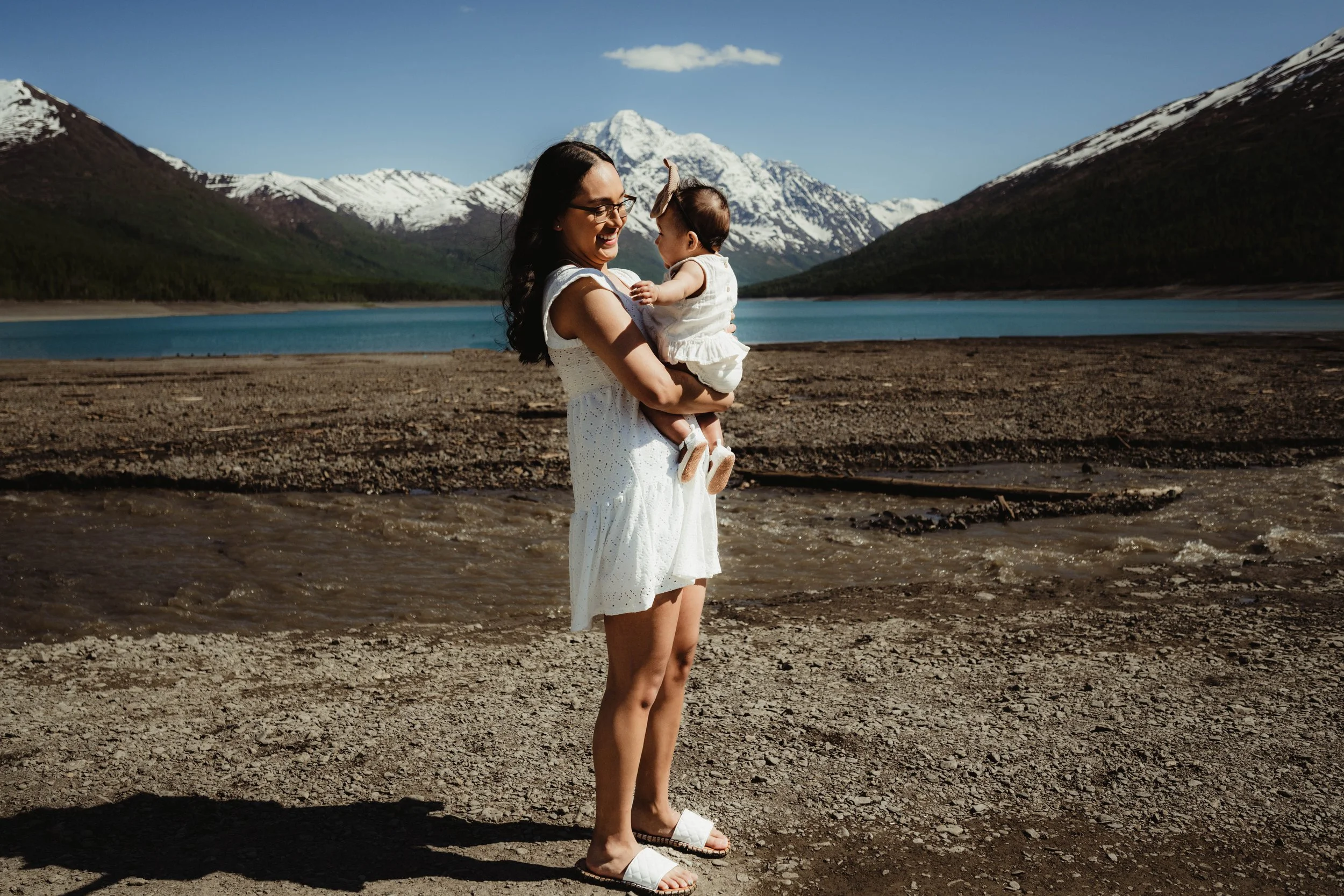 A woman holding a baby girl by a lake surrounded by mountains with snow, during daytime.