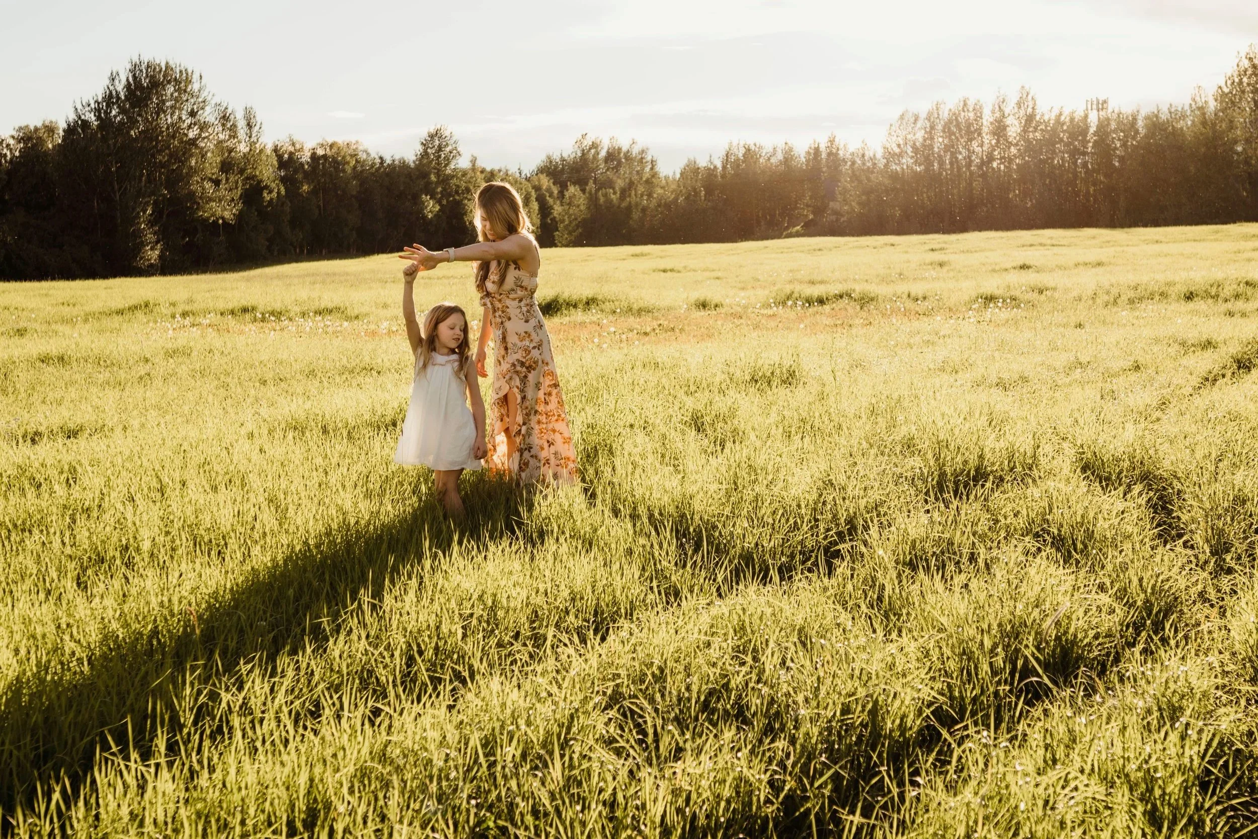 A woman and a young girl dancing together in a green field during sunset, with trees in the background.