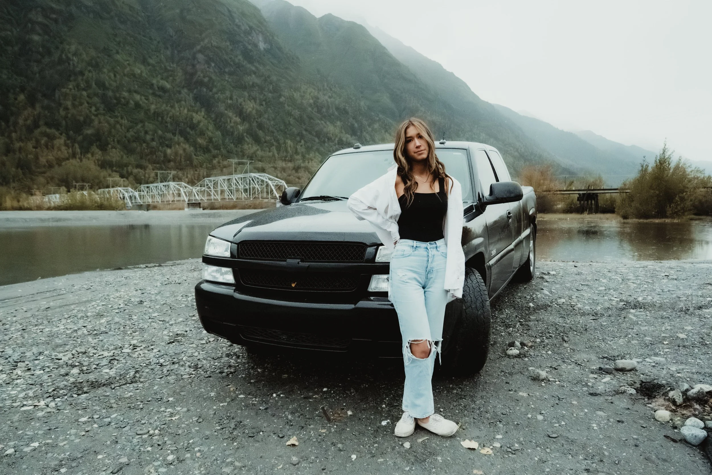 A young woman in ripped jeans, black top, white jacket, and white sneakers stands next to a black pickup truck on a gravel surface with a river, mountains, and a bridge in the background.
