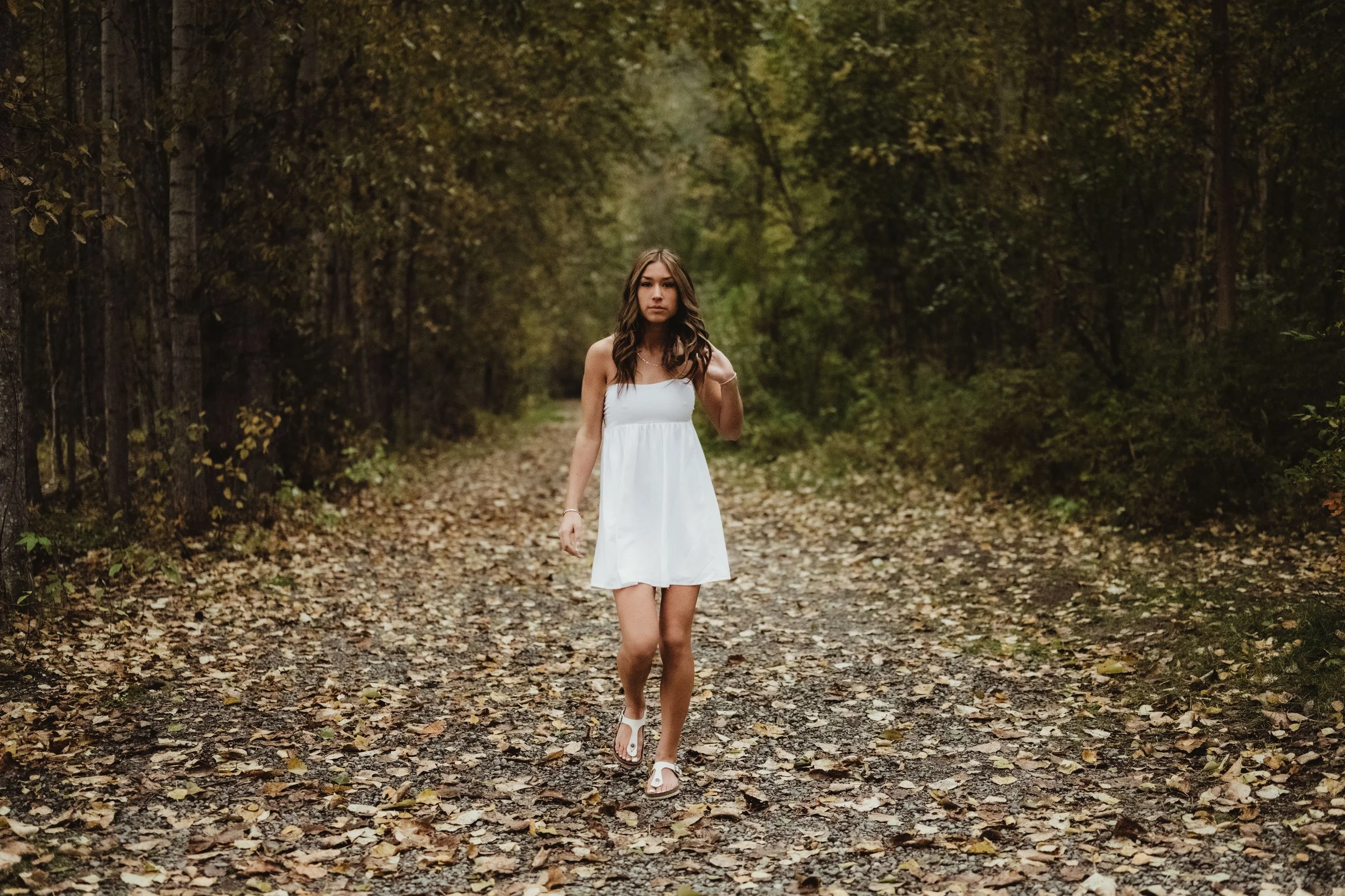A young woman in a white dress walking along a leaf-covered forest path surrounded by trees with green and brown foliage.