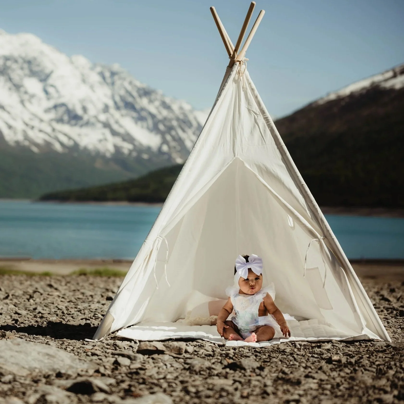 A baby sitting inside a small white tent on a rocky outdoor area with a mountain and lake in the background.