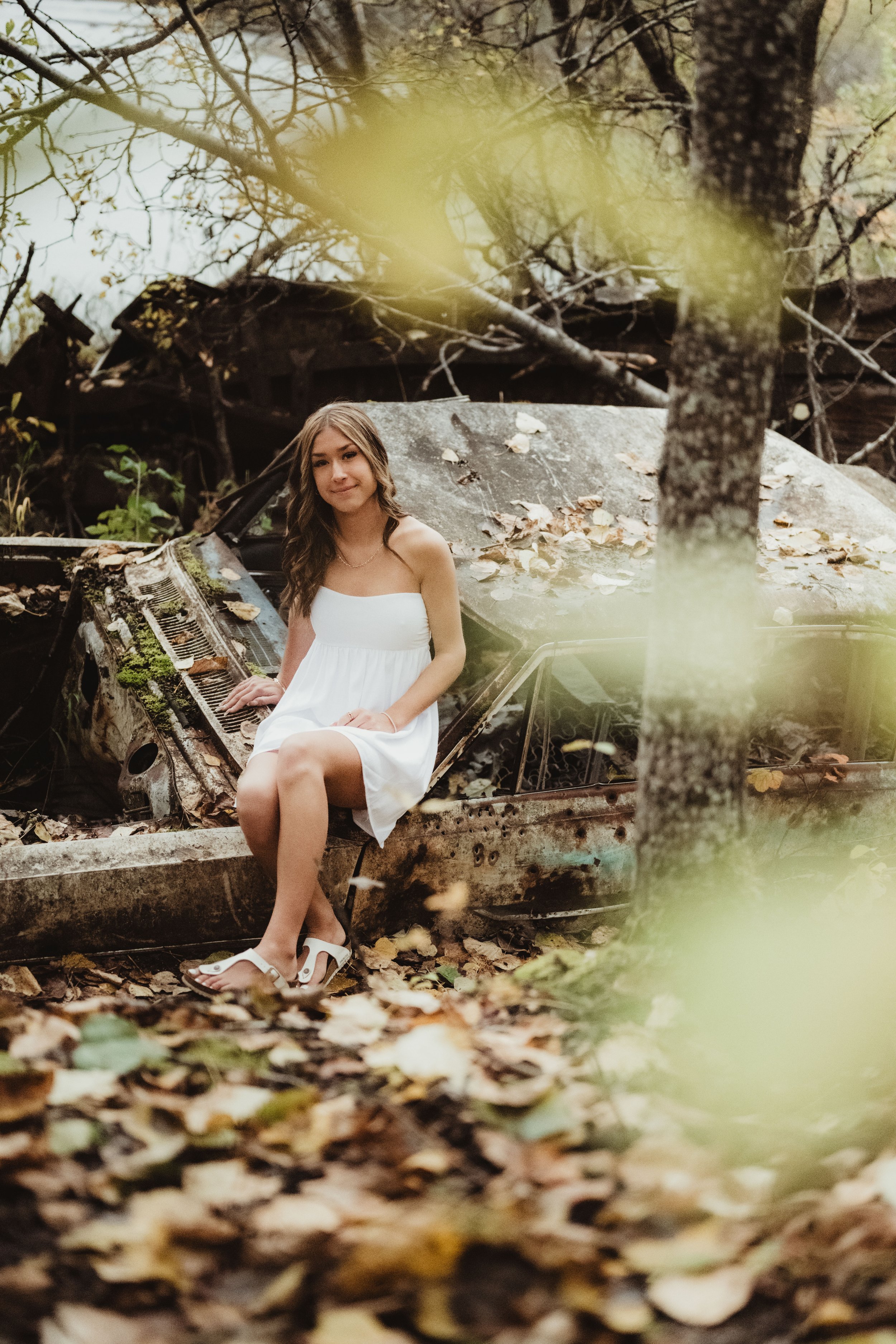 A young woman in a white dress sitting on the hood of an abandoned, rusted car surrounded by fallen leaves and trees.