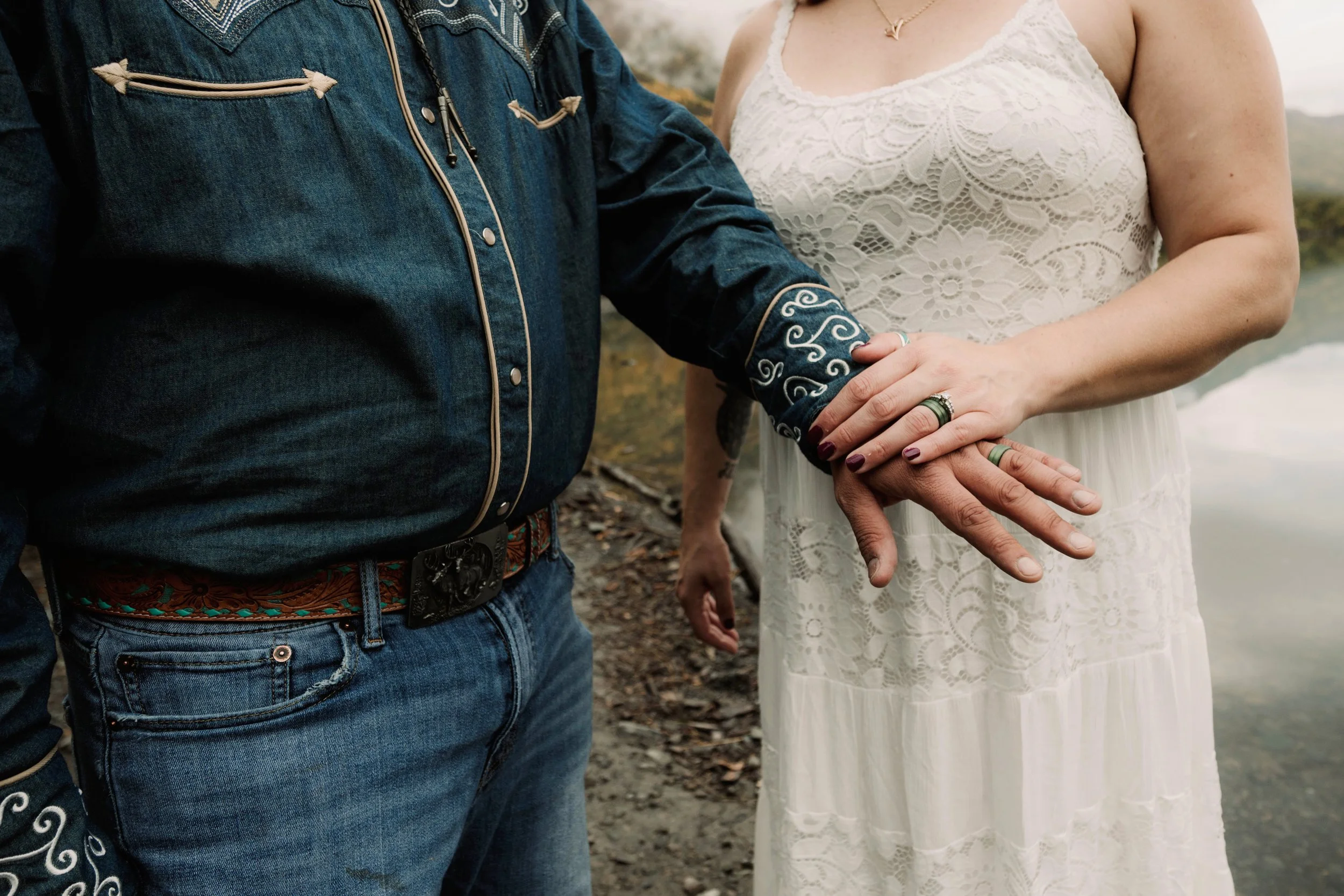 A couple holding hands by a body of water, one wearing a white lace dress and the other in Western-style denim shirt and jeans.