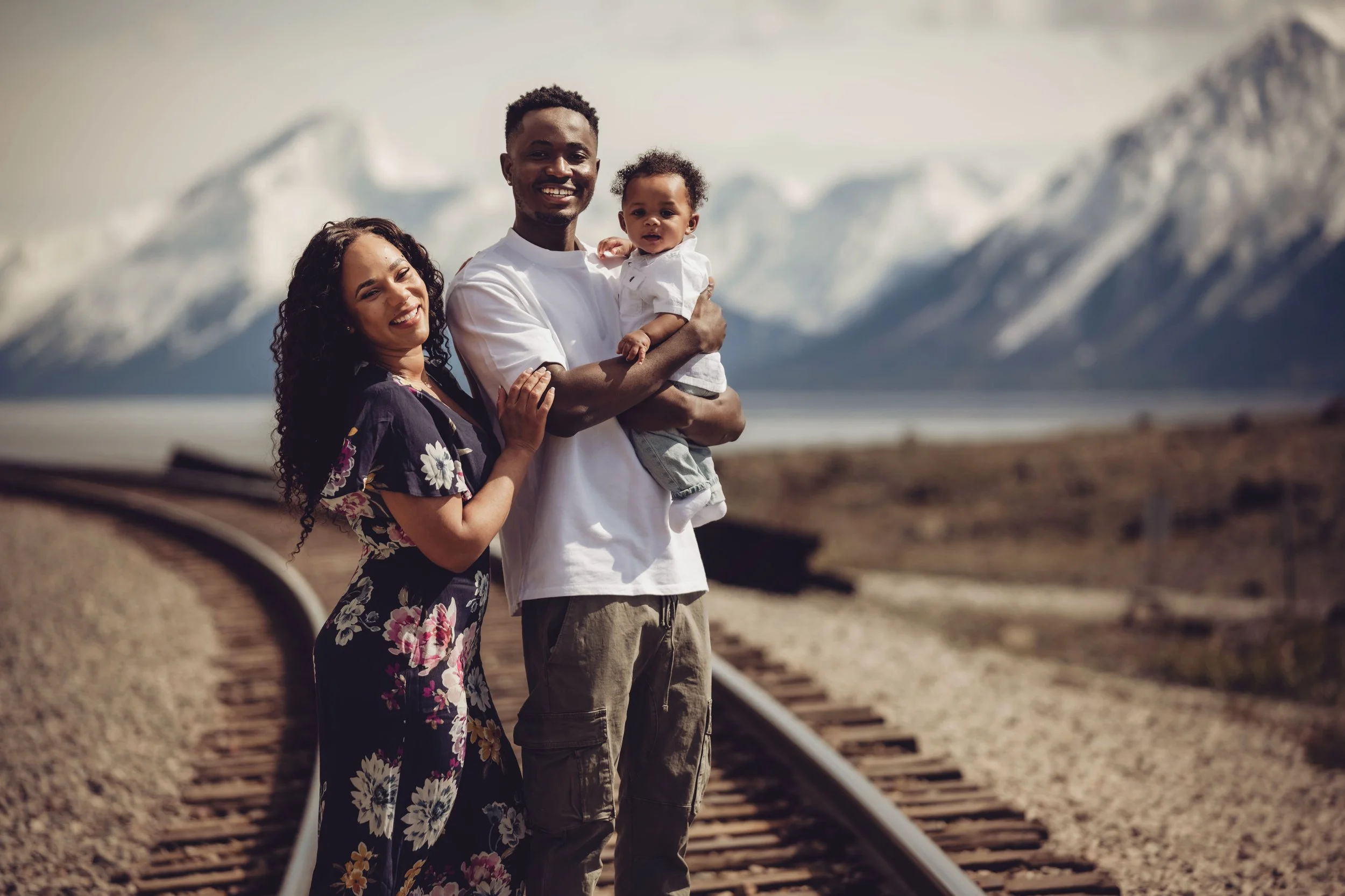 A happy family of three standing on train tracks in front of snow-capped mountains, with a lake in the background. The woman has curly hair and is wearing a floral dress, the man is wearing a white t-shirt, and the child is being held by the man wearing a white shirt.