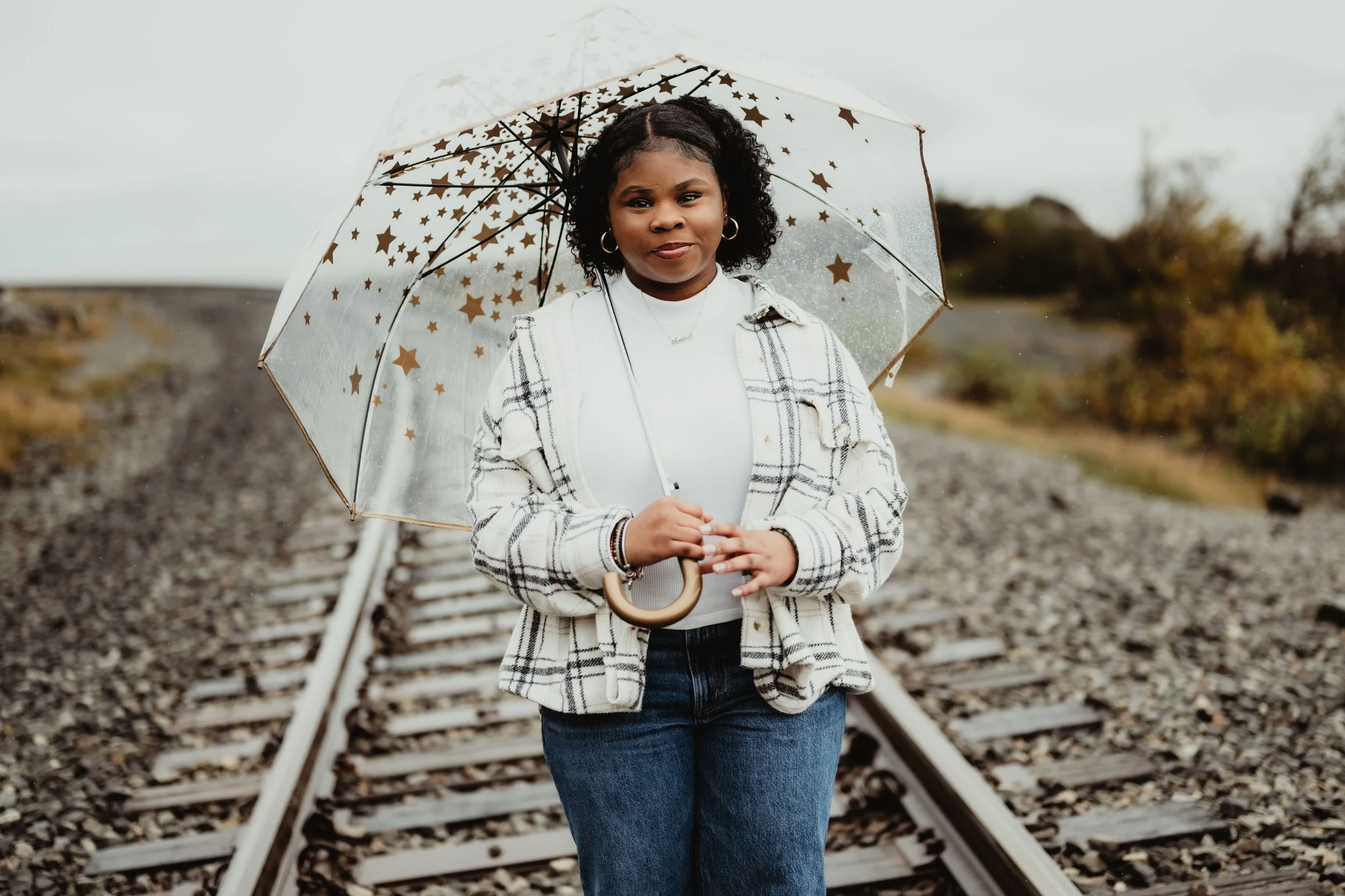 A woman holding a umbrella standing on train tracks outdoors, wearing a white outfit with a plaid jacket, with cloudy skies and trees in the background.