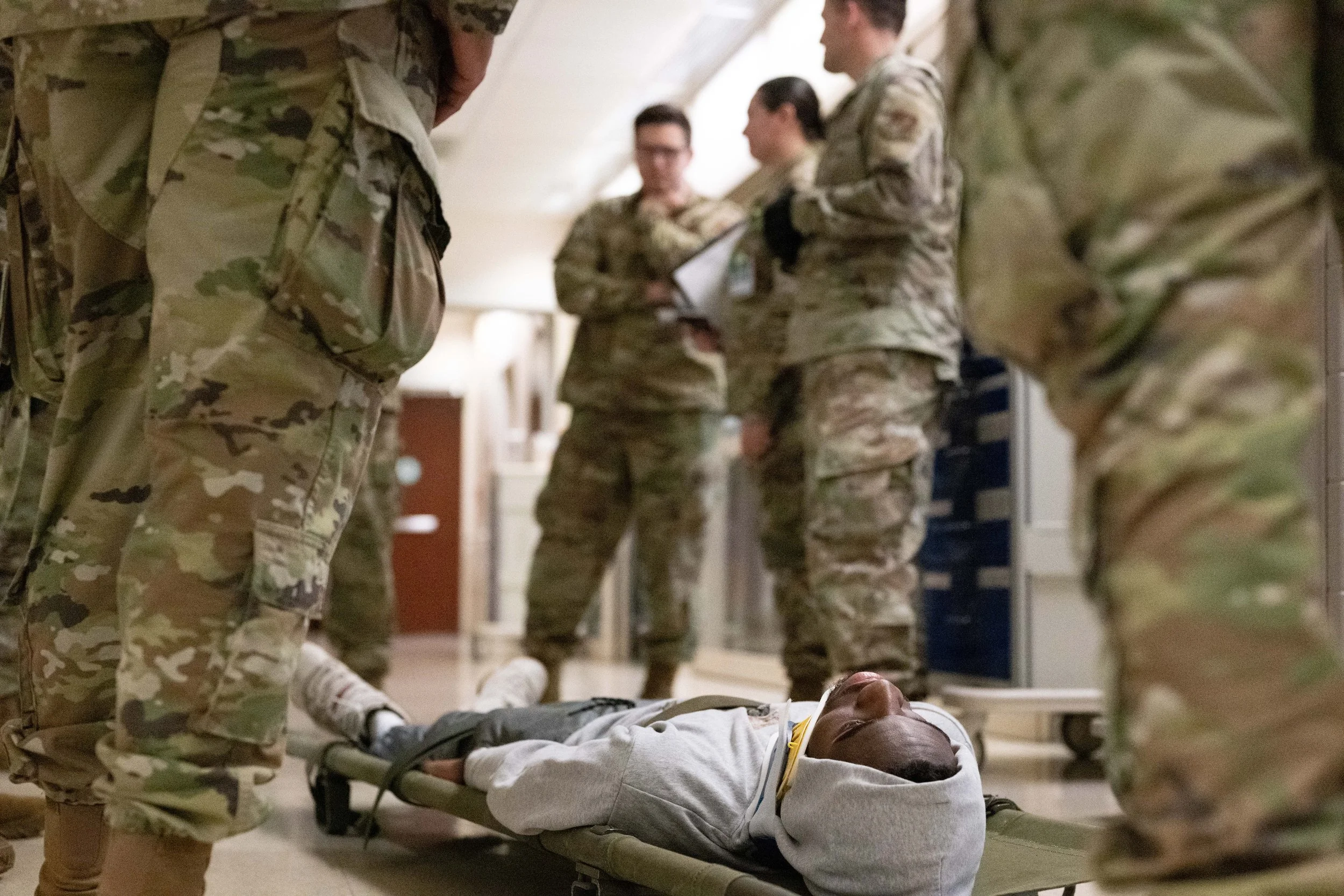 Military personnel attend to an injured person on a stretcher in a hospital or medical facility.