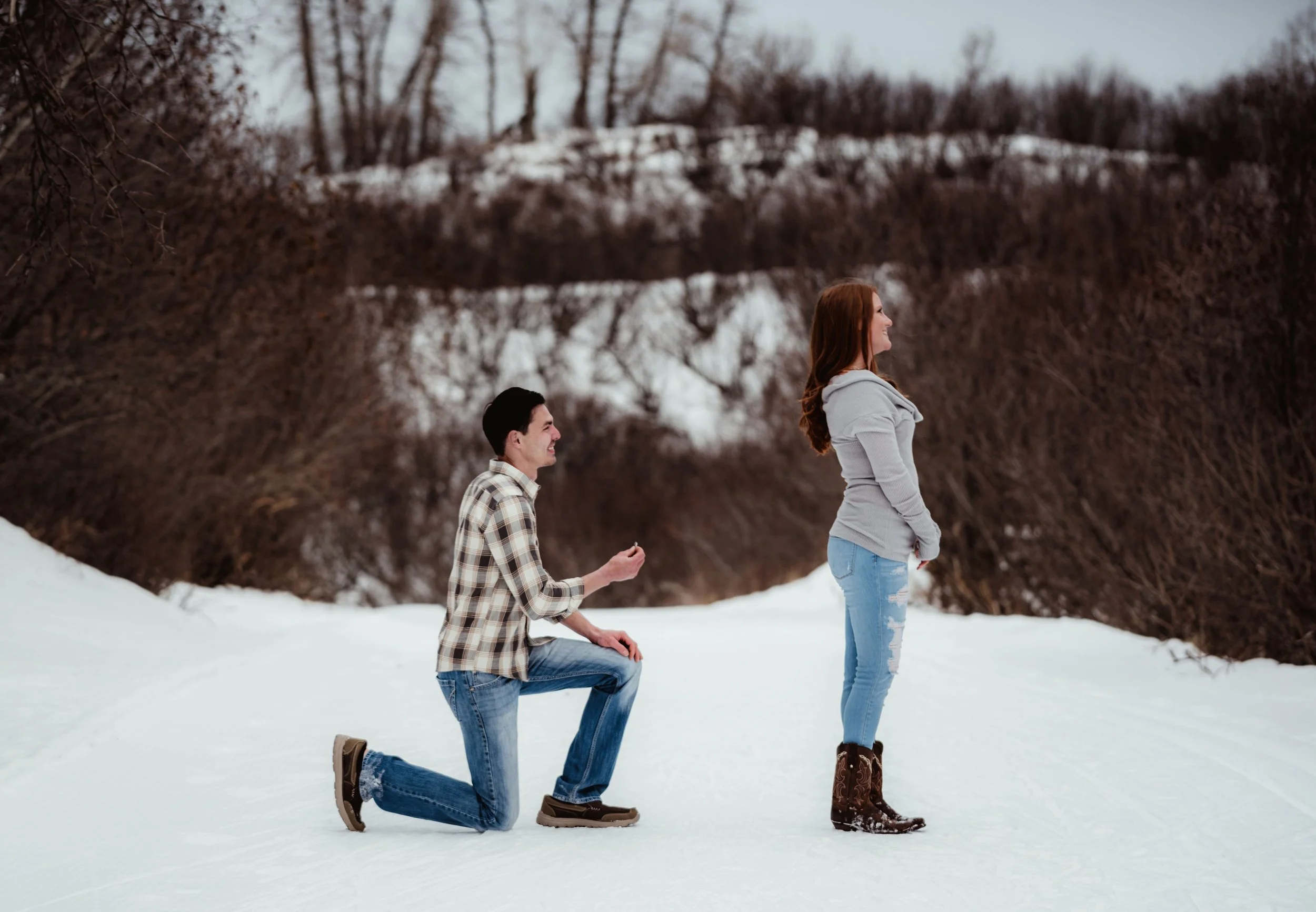 A man on one knee proposing to a woman on a snowy trail outdoors during winter, with a background of snow-covered hills and leafless trees.