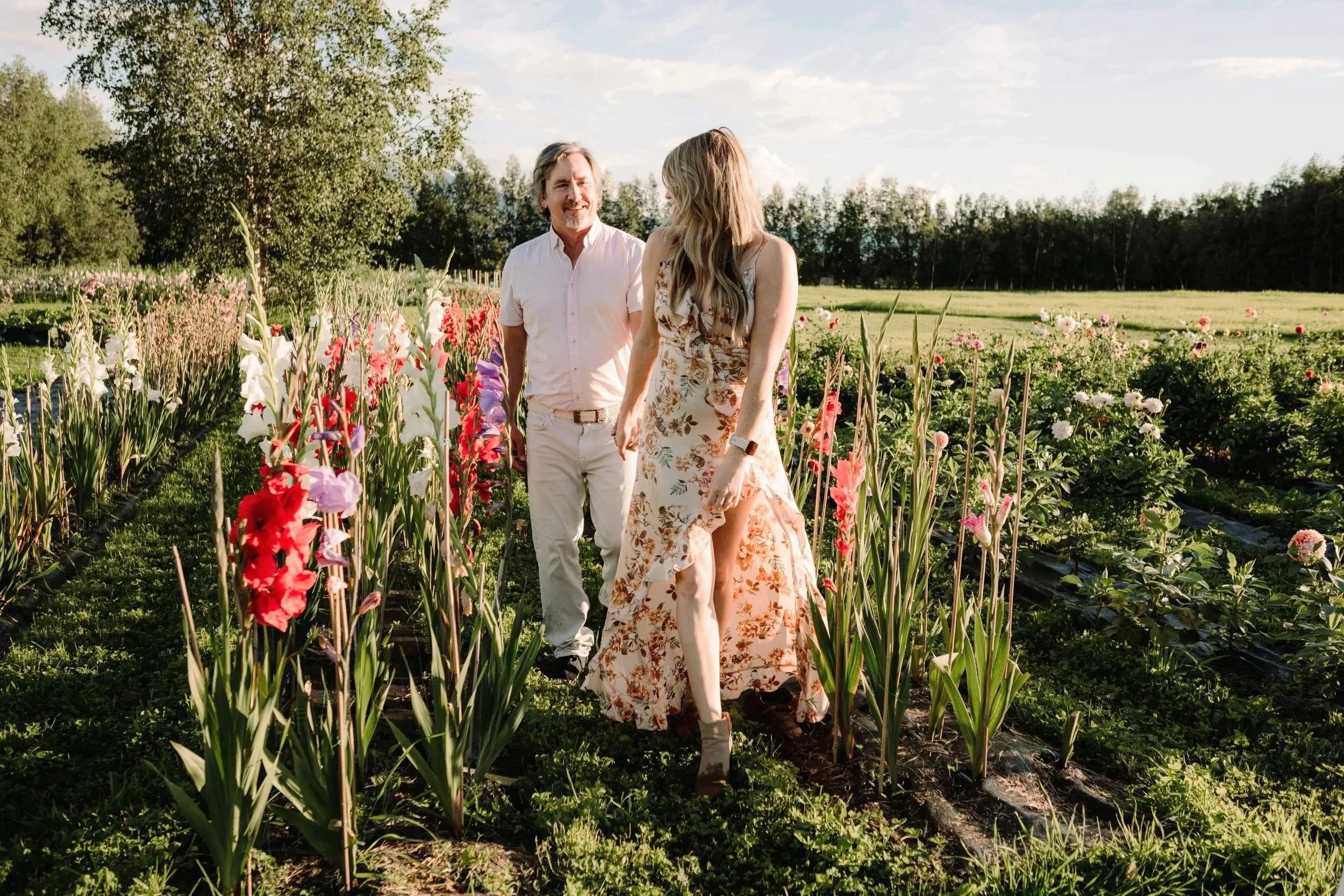 A man and woman walking through a flower garden on a sunny day, with the woman wearing a floral dress and the man in a white shirt and light-colored pants.