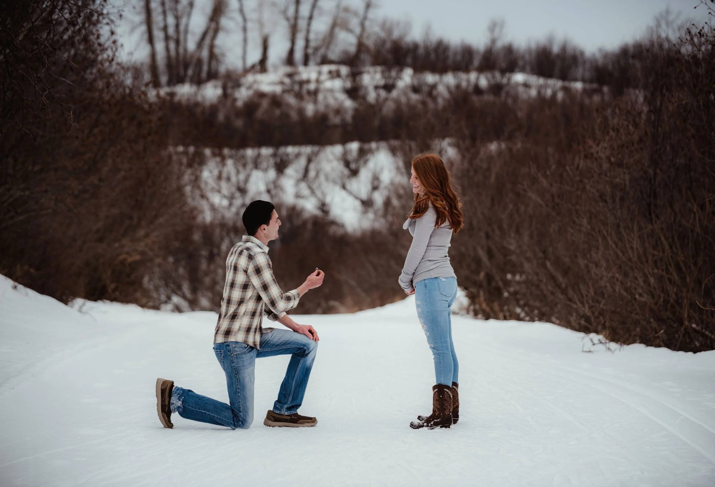 A couple with a man kneeling and proposing to a woman in a snowy outdoor setting during winter.