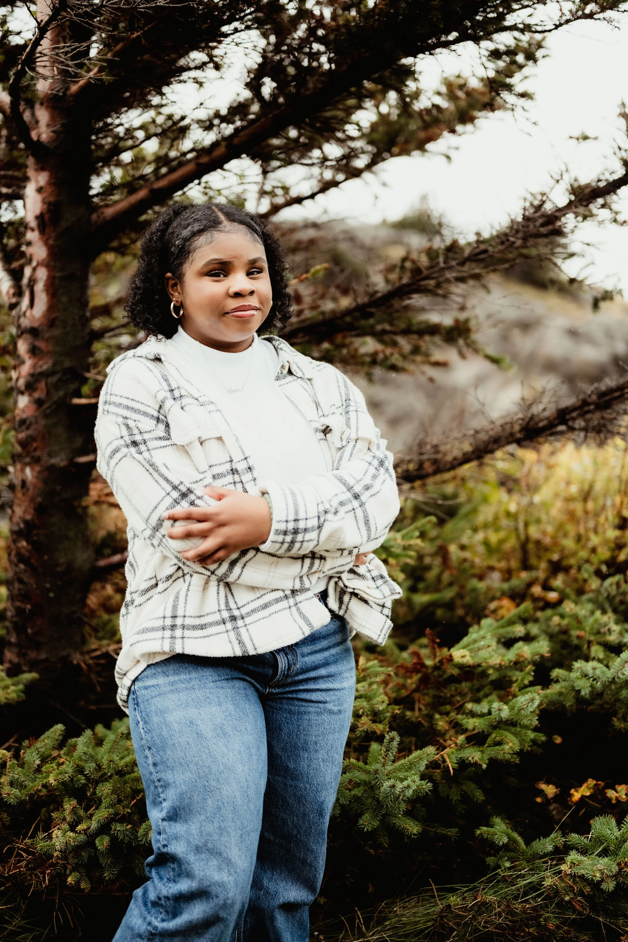 A young woman with curly hair, wearing a white plaid jacket and blue jeans, stands outdoors in front of a tree with her arms crossed, smiling slightly.
