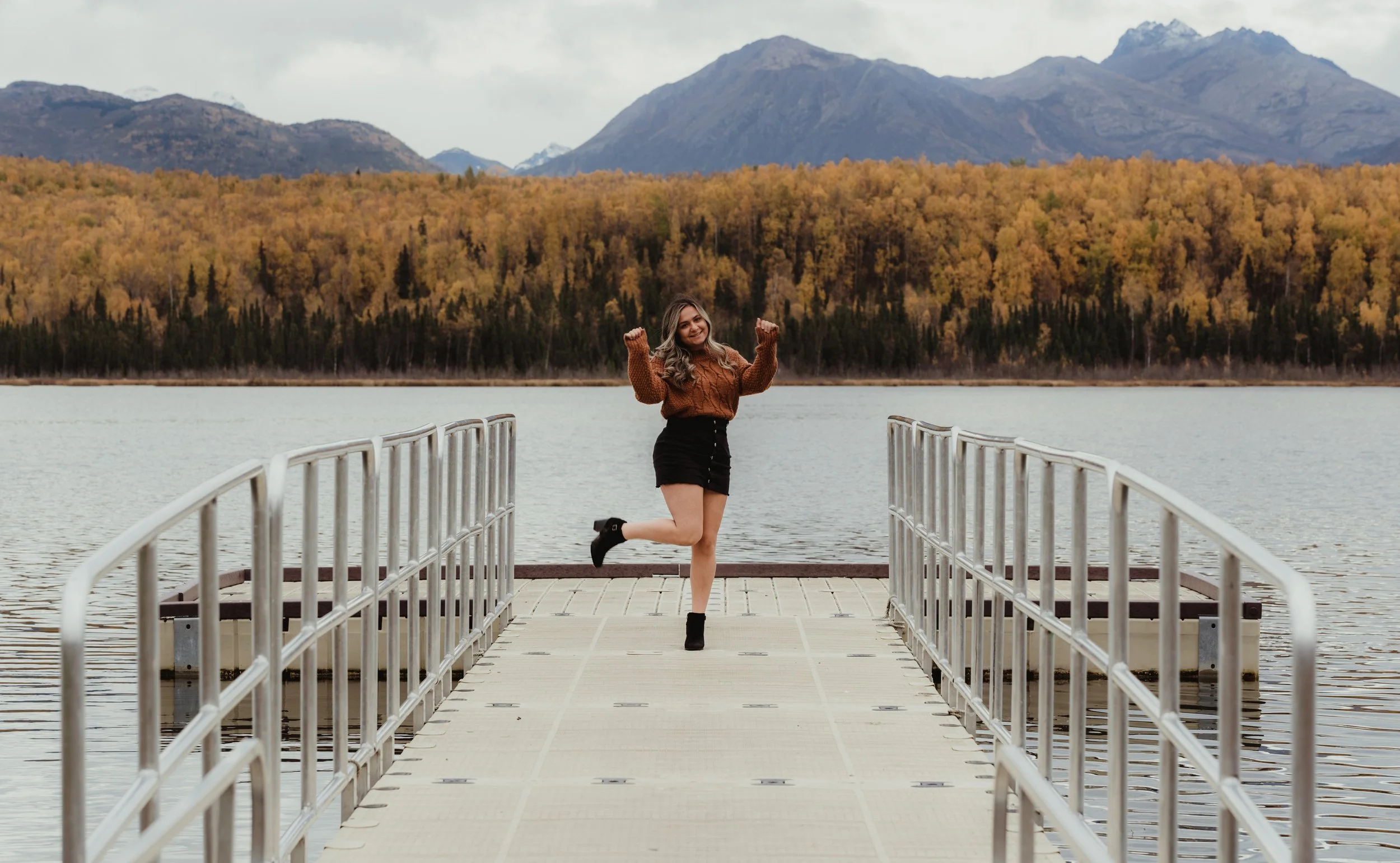 A woman standing on a dock by a lake, with mountains and autumn trees in the background. She is smiling, wearing a brown sweater, black shorts, and black boots, and is posing with one leg lifted and arms raised.