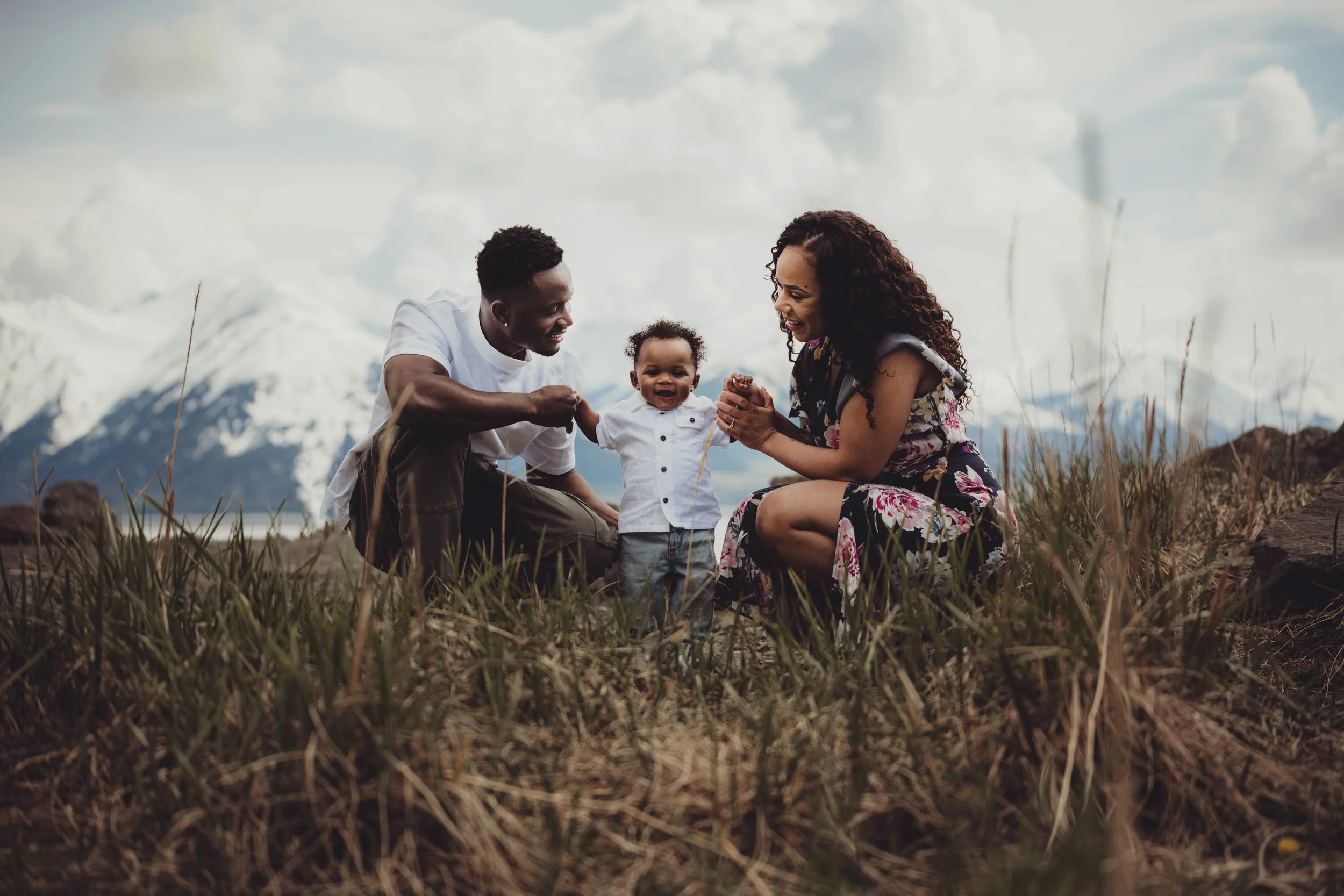 A family of three, including a woman, a man, and a young child, outdoors on a grassy field with snow-capped mountains in the background. The family members are smiling and playing together.