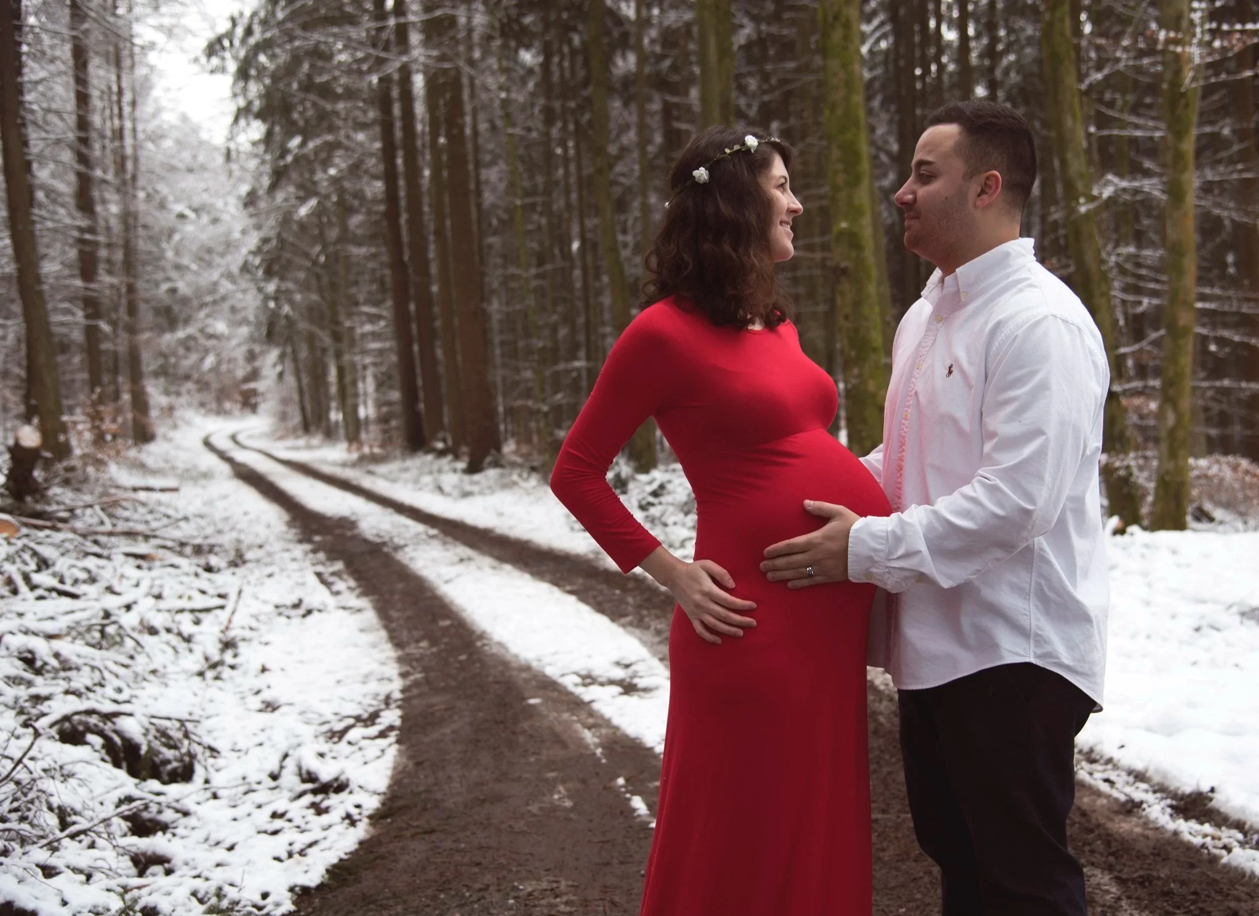 A pregnant woman in a red dress and a man in a white shirt standing in a snowy forest, facing each other and touching her belly