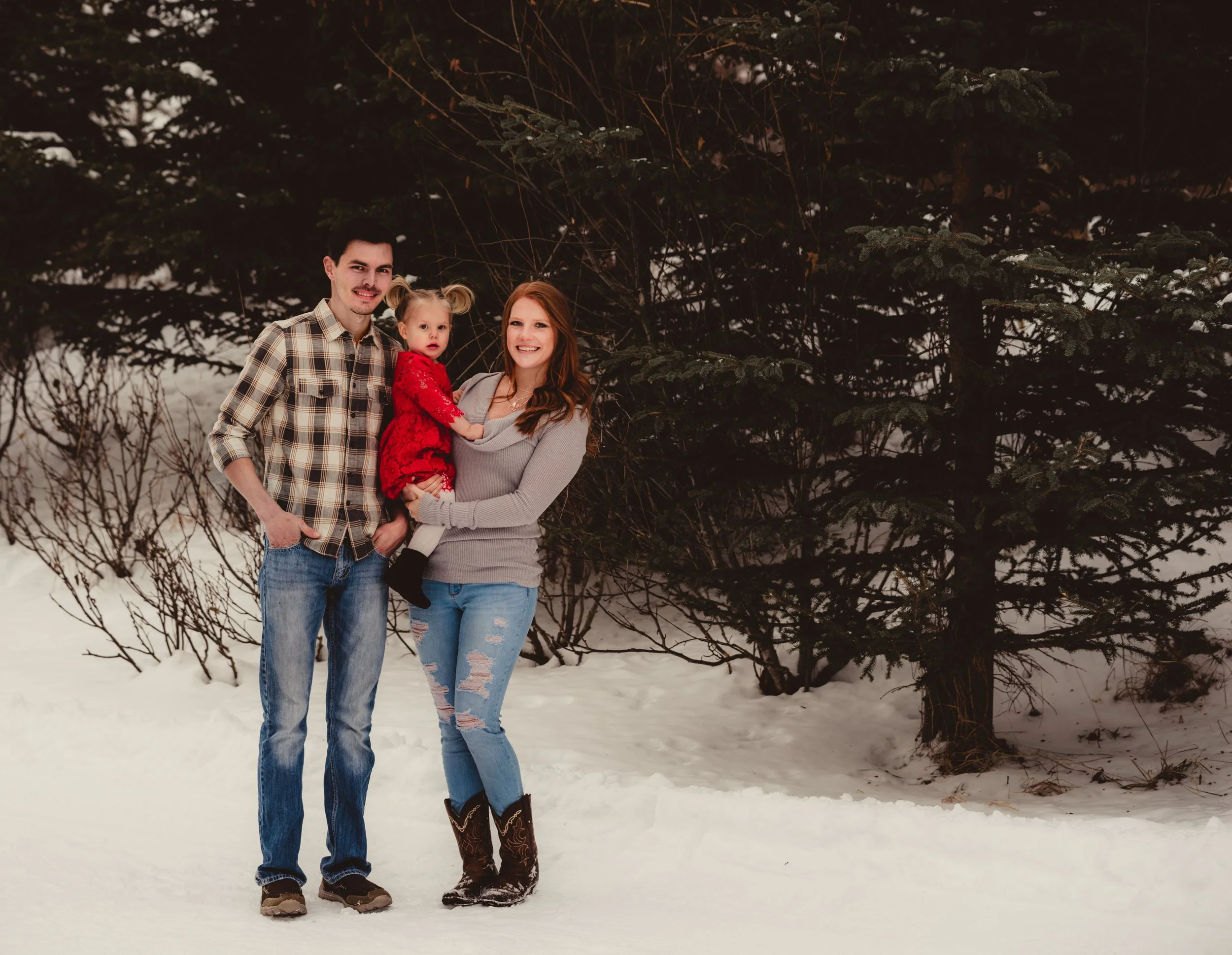 A family of three standing outdoors in snow with evergreen trees in the background during winter, smiling at the camera.