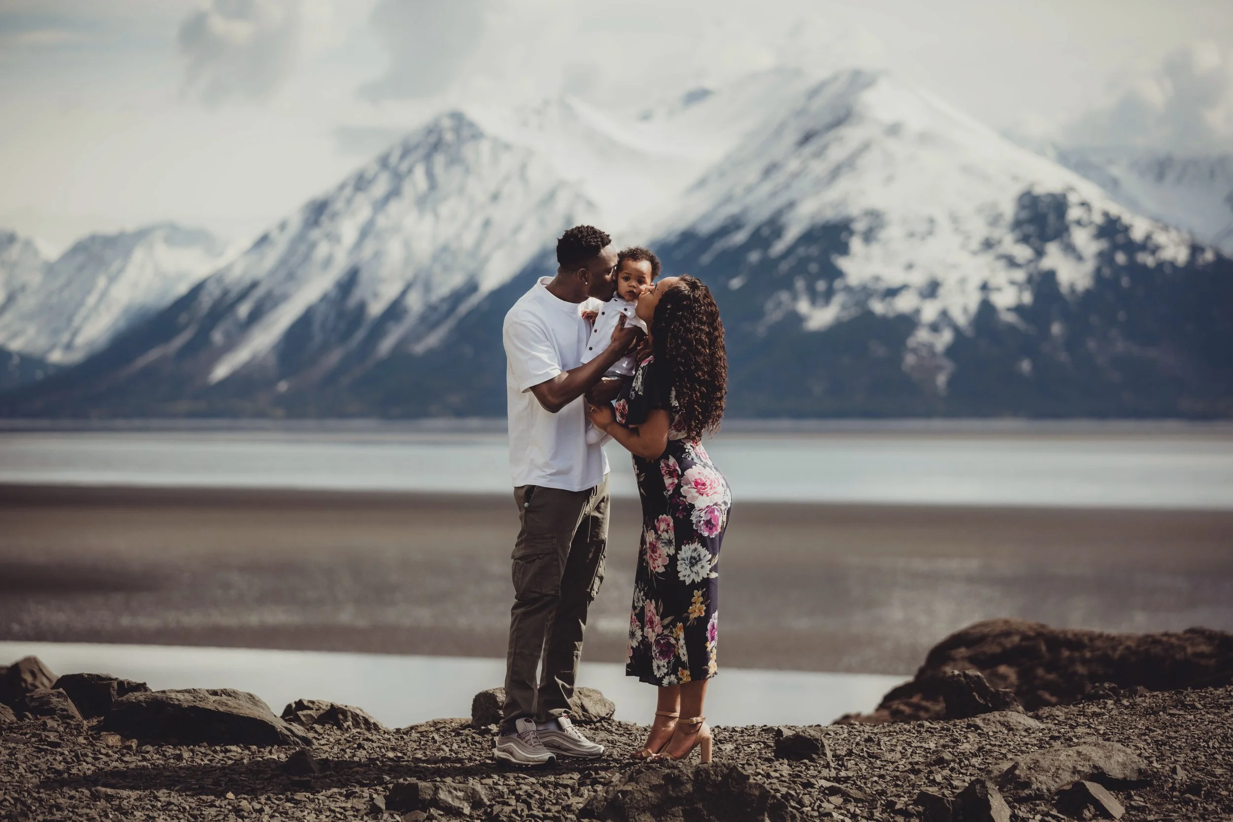 A family of three standing on rocks near a lake with snow-capped mountains in the background. The father is holding their young son, and the mother is kissing him.