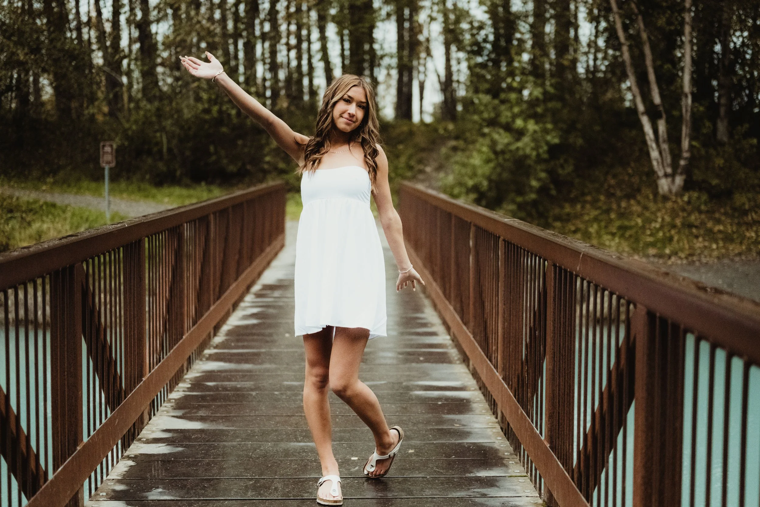 Young woman in a white dress and flip-flops standing on a wooden bridge surrounded by trees, with one arm raised and a smile.