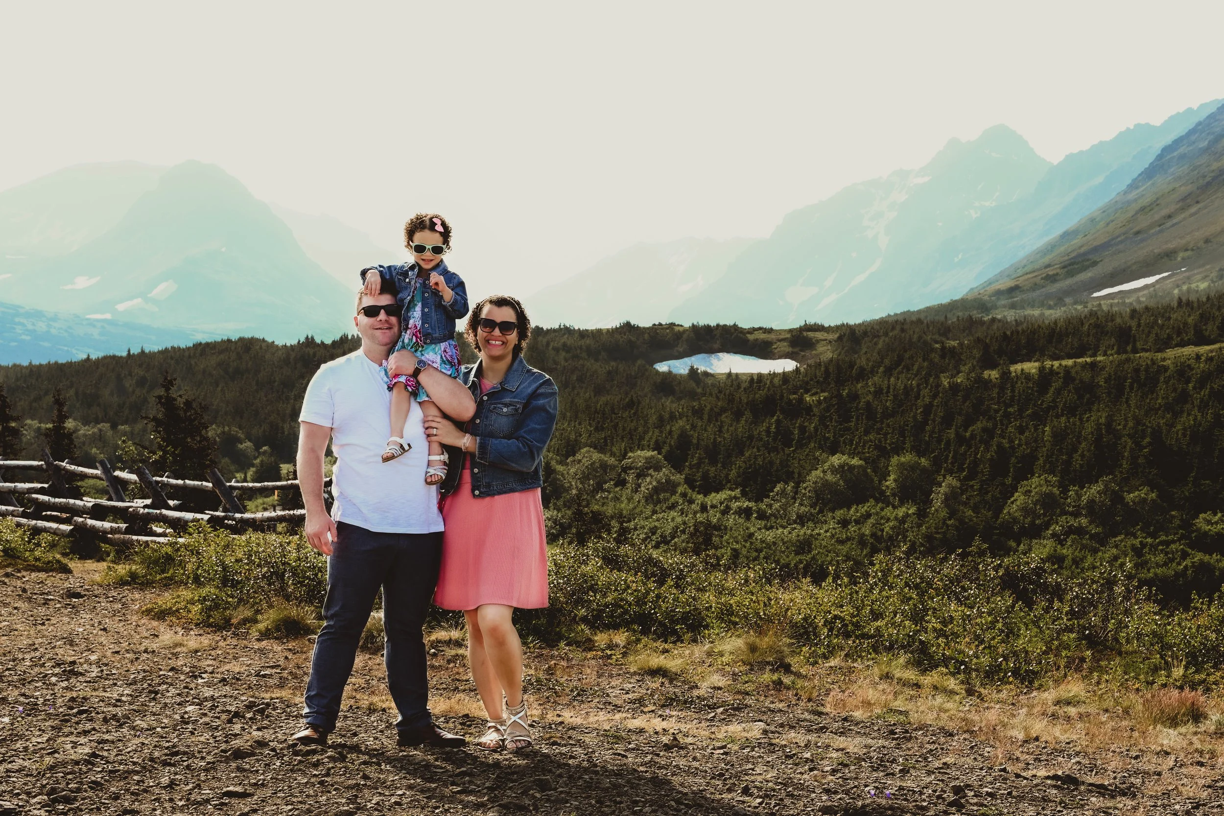 A family of three, with a man, woman, and young girl, standing outdoors on a dirt path in front of mountains and forest, smiling at the camera.