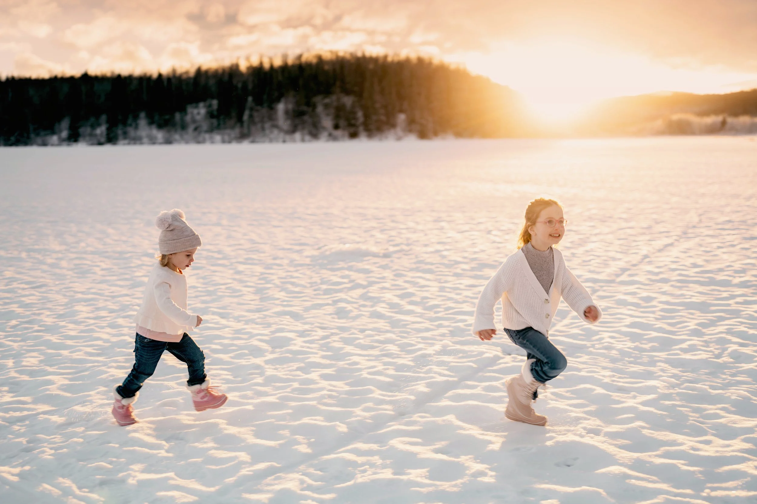Two young girls running and playing in a snow-covered field during sunset, with a forest in the background.