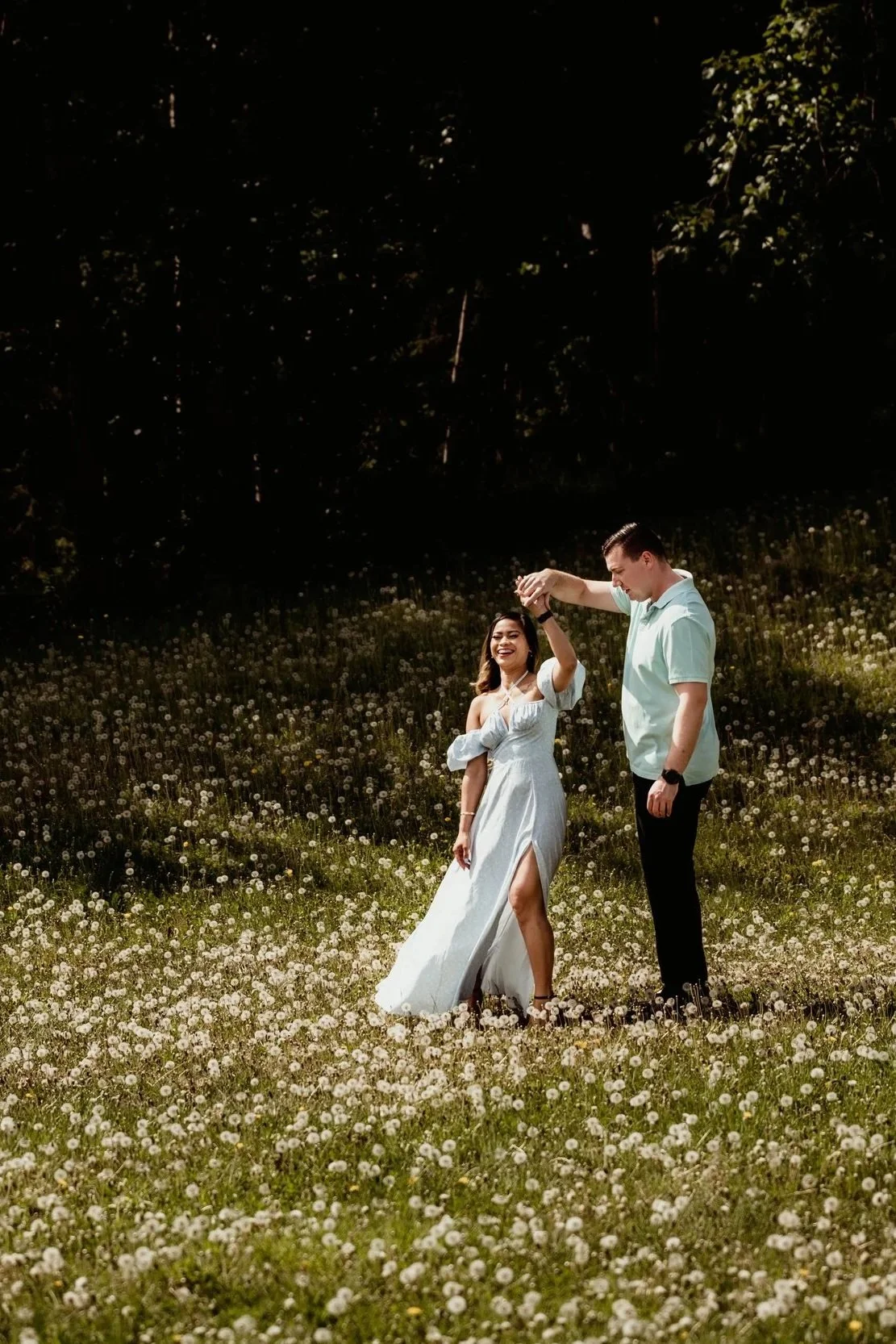 A man and woman dancing in a field full of small white flowers, with trees in the background, during daylight.
