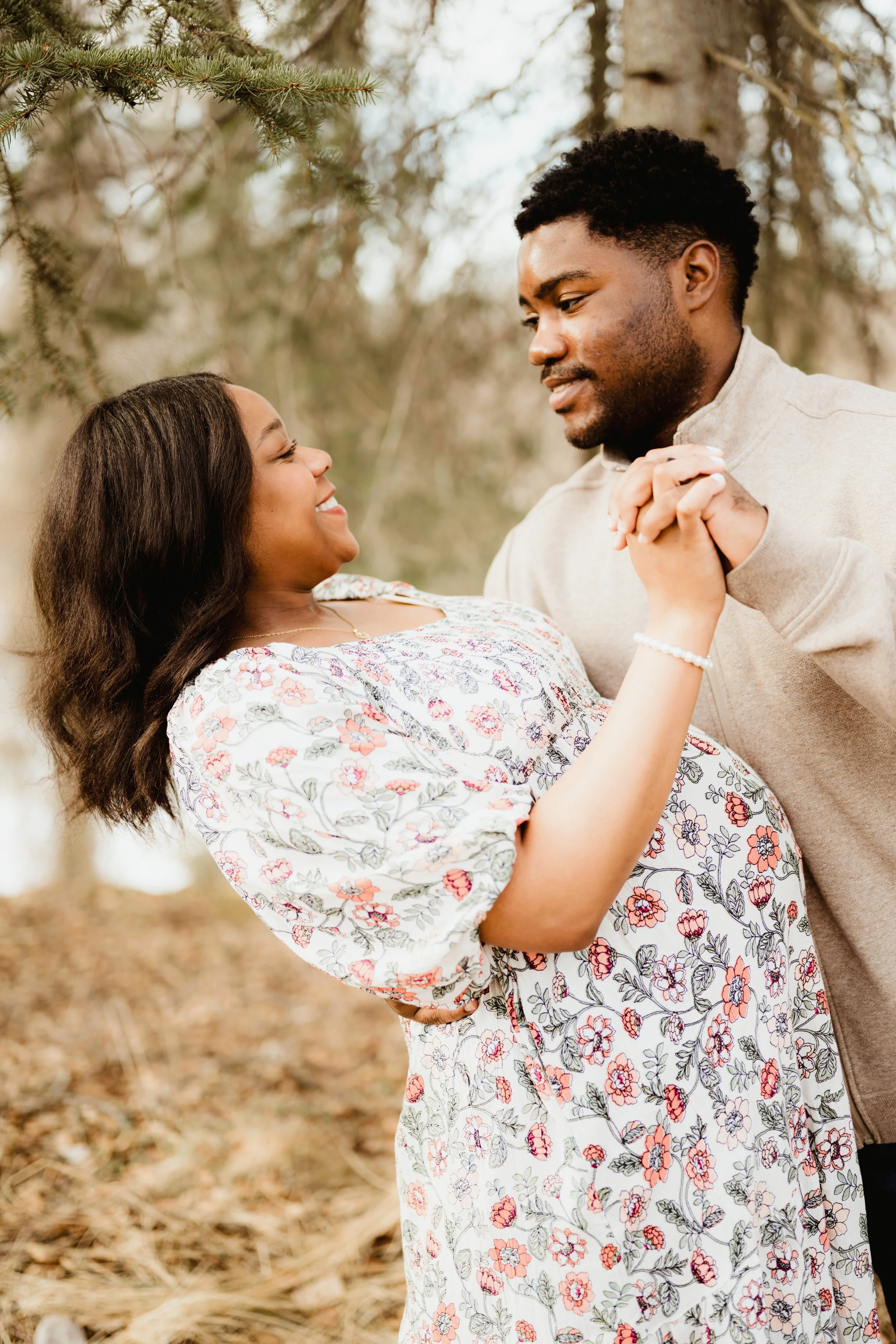 A pregnant woman and a man dance outdoors in a natural setting with trees, holding hands and smiling at each other.