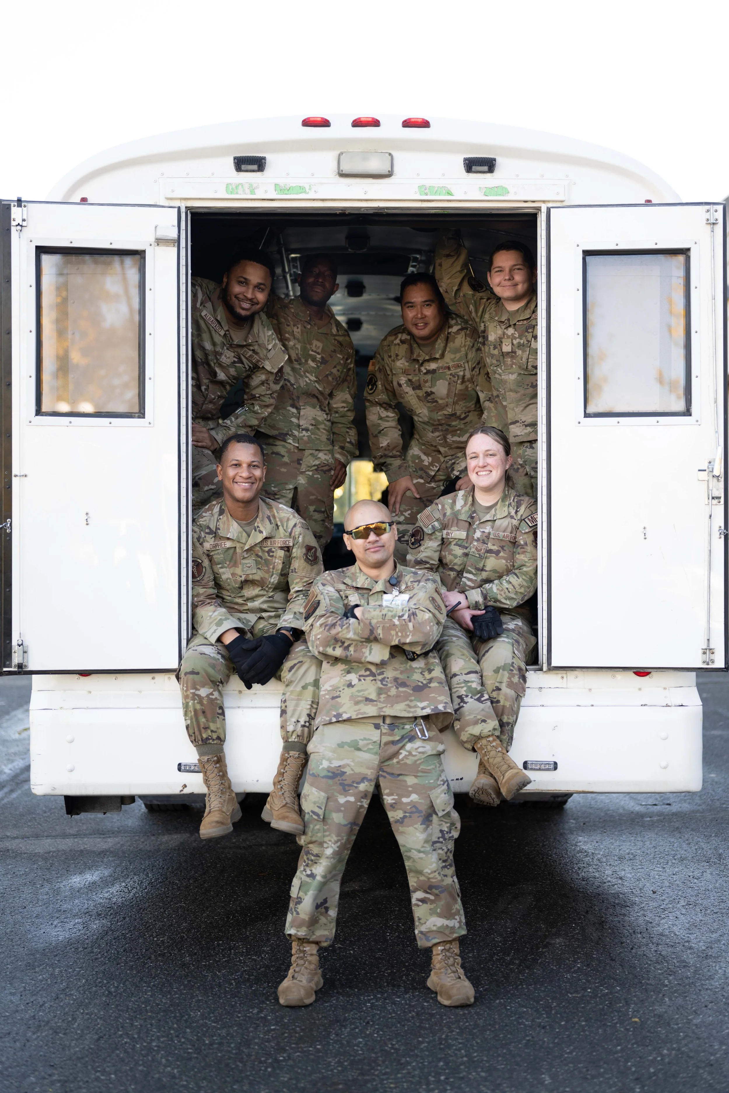 Group of seven soldiers in camouflage uniforms posing in front of a white military vehicle, some sitting and some standing inside.