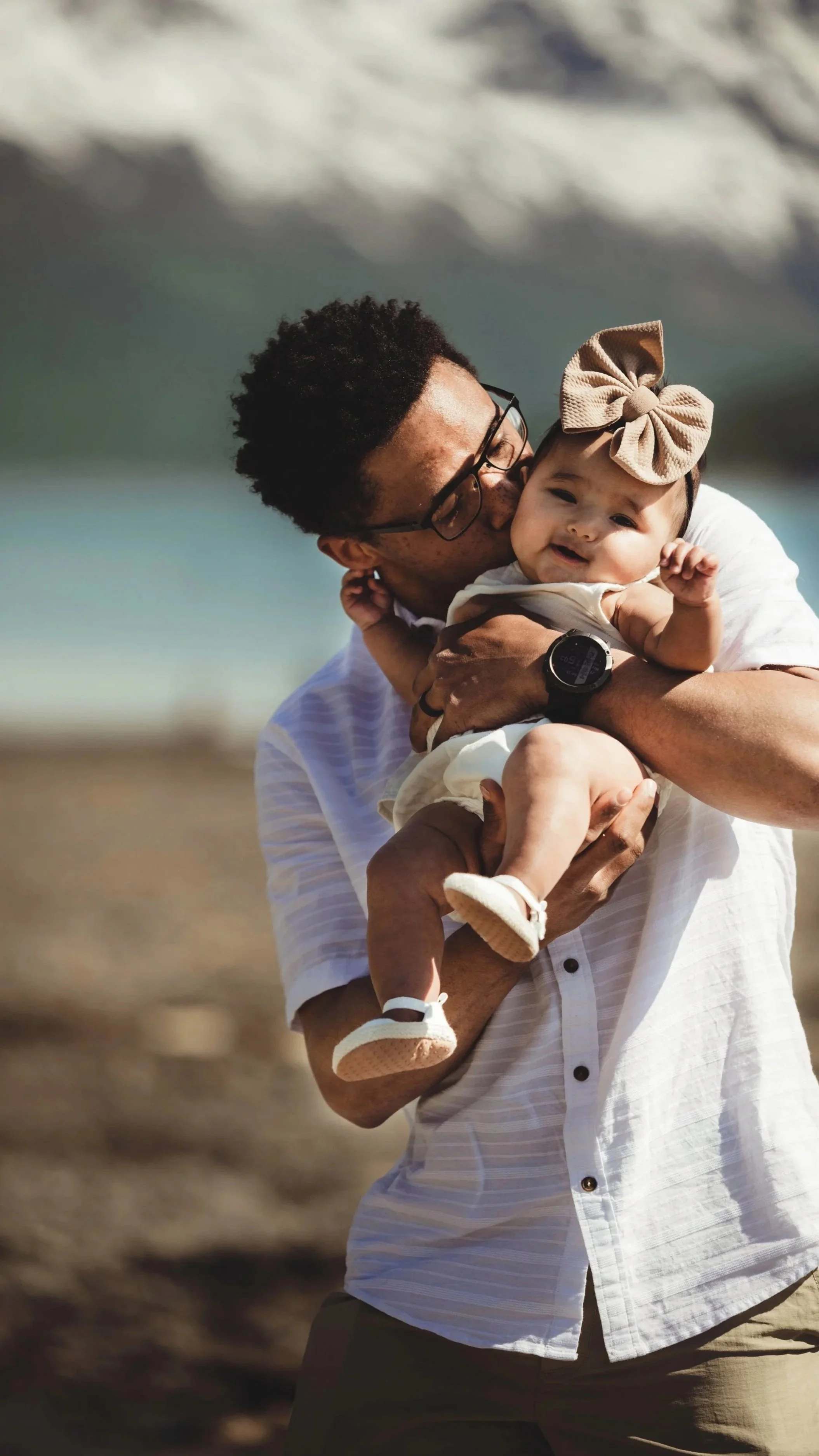 A man holding and kissing a smiling baby girl on the beach with ocean waves in the background.