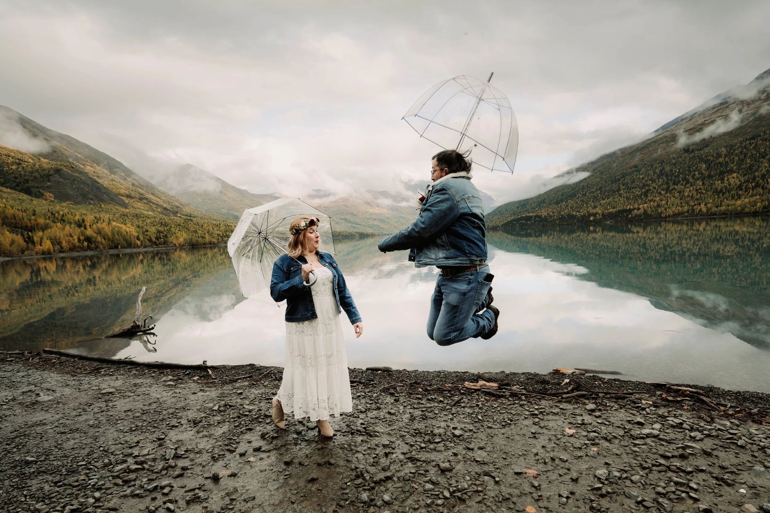 A couple with umbrellas near a lake, one person is jumping while the other stands on the rocky shore, surrounded by mountains and cloudy sky.