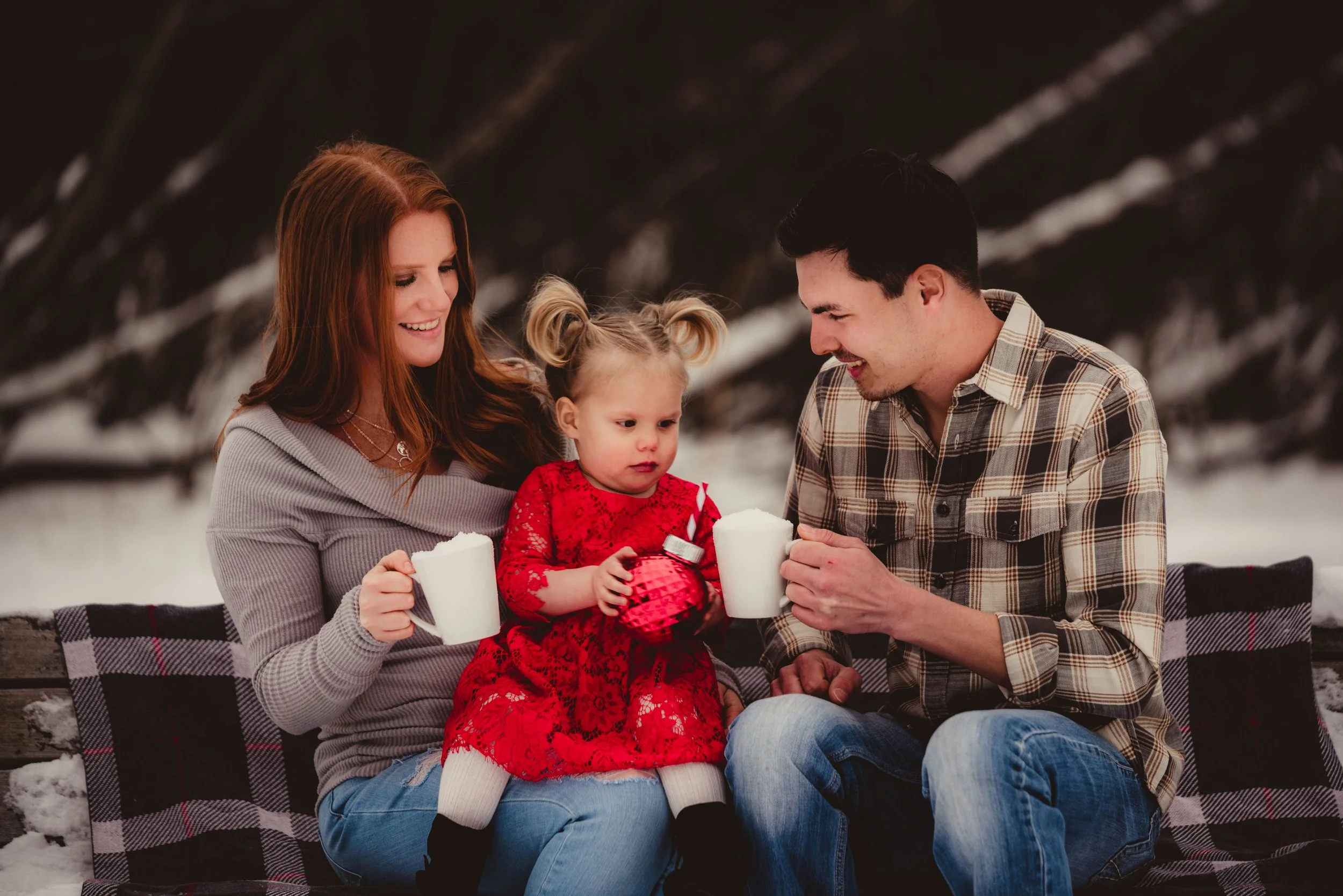 A family of three sitting on a park bench outdoors in winter. The mother with red hair is smiling while holding a mug, the father with dark hair is smiling and holding a mug, and their young daughter in a red dress is holding a Christmas ornament and looking at it with a serious expression. Snow is visible on the ground and trees are in the background.