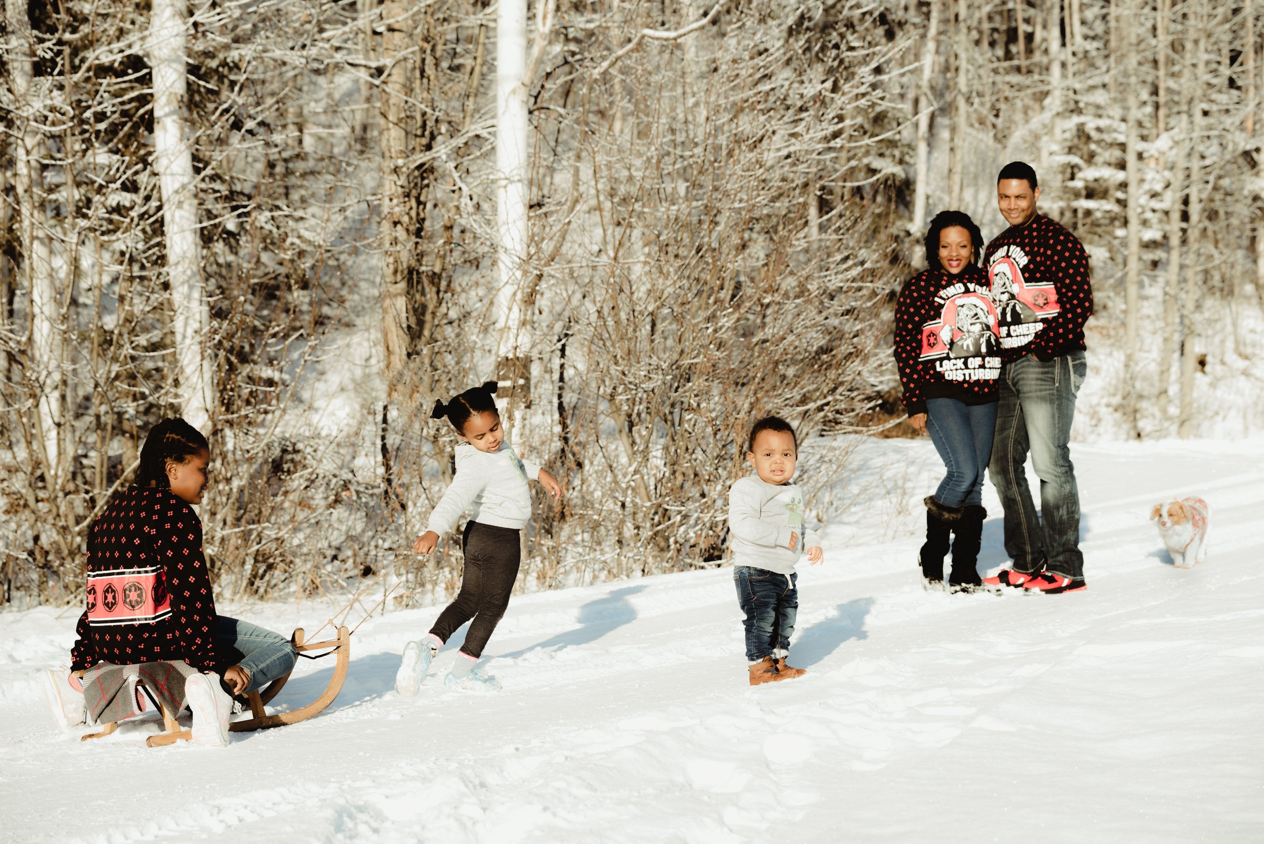 Family playing in the snow: two children in the foreground, one sitting on a sled, another running, and an adult couple standing with a small dog, with snow-covered trees in the background.