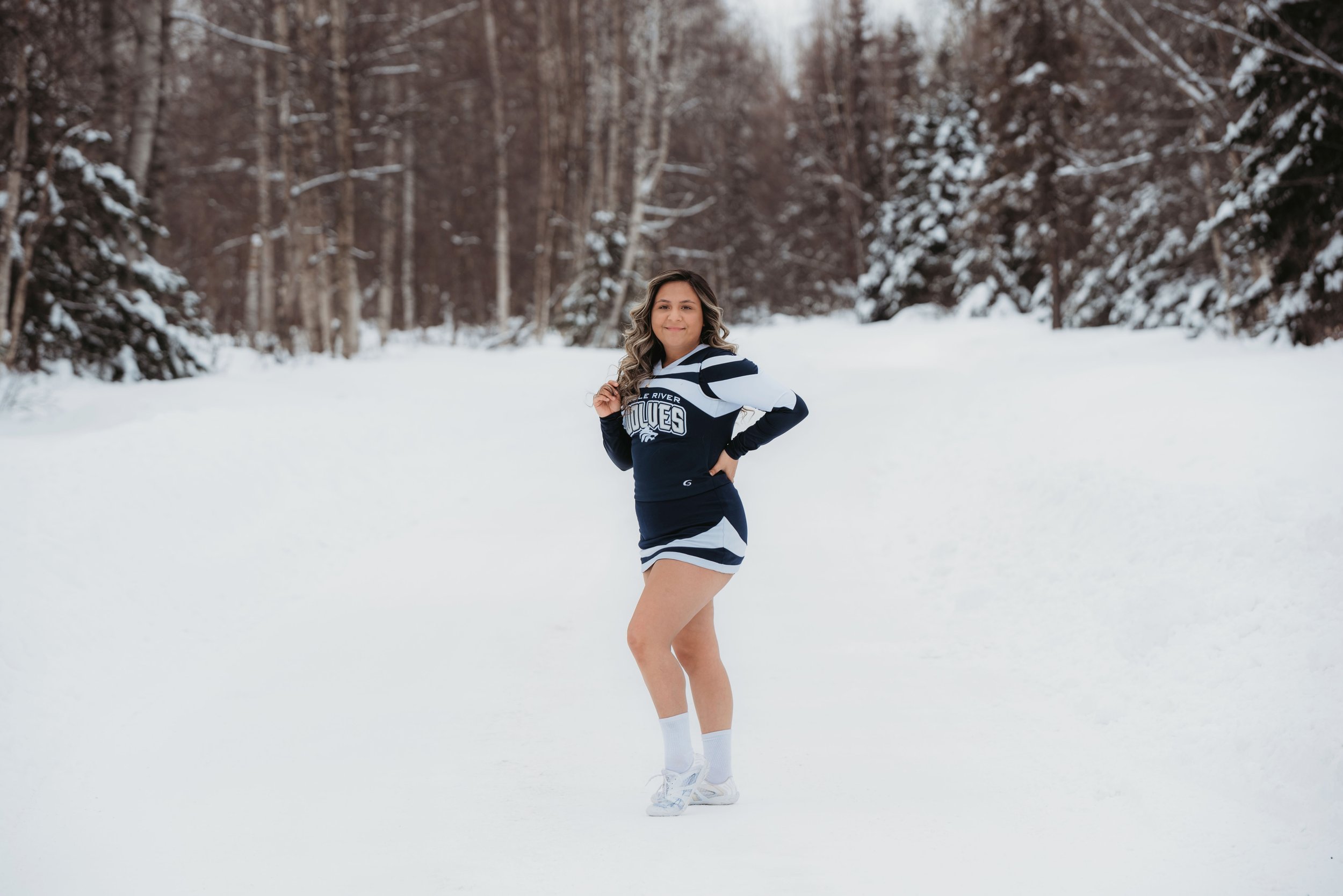 A woman stands in a snow-covered forest, wearing a navy and white sports outfit with the word "BULLIES" on her top, white shoes, and socks, smiling at the camera.