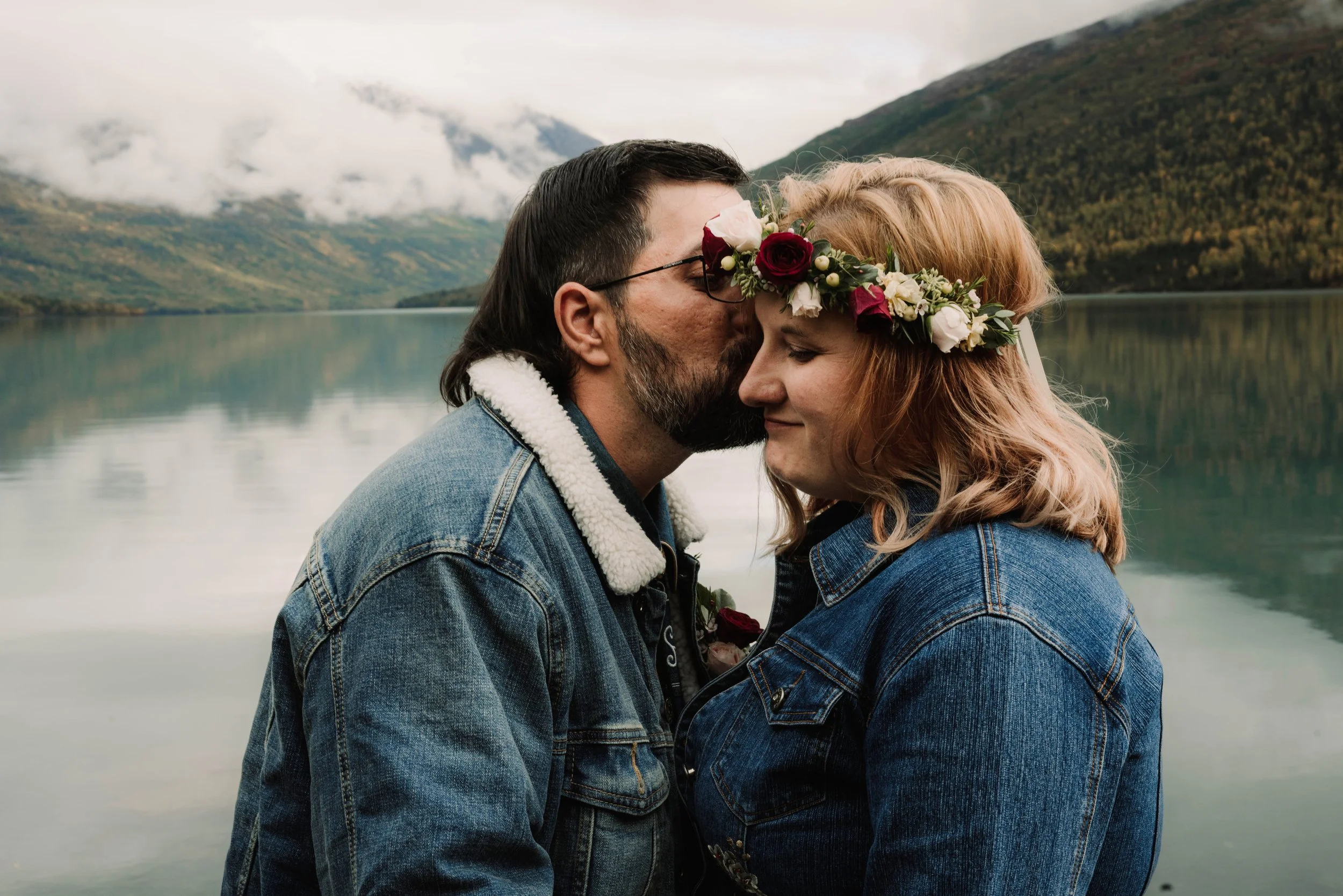 A couple in denim jackets sharing a kiss by a lake with mountains in the background, woman wearing a floral crown.